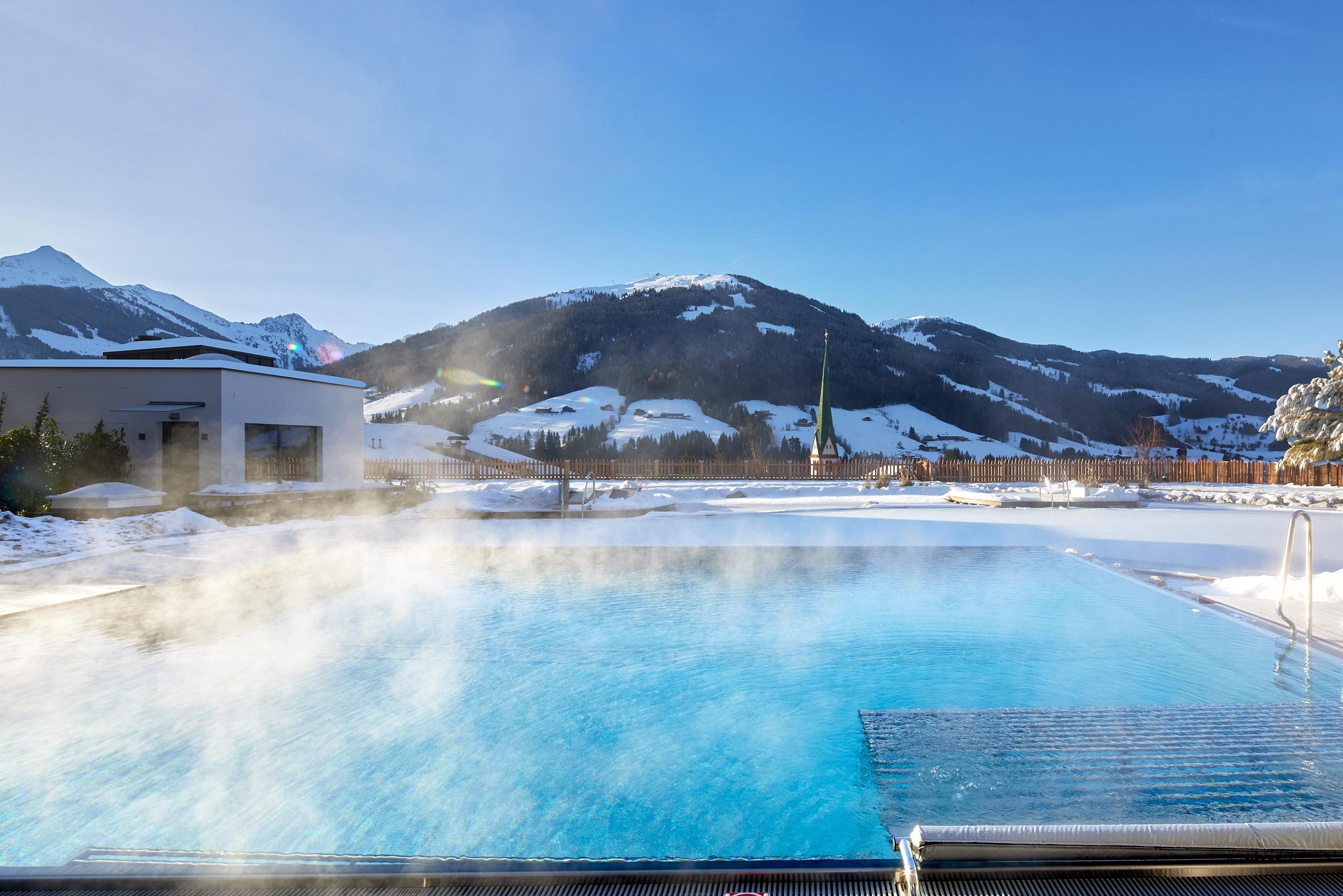 A beautiful pool in the snow with steaming water surface. In the background, majestic mountains and a clear blue sky can be seen.