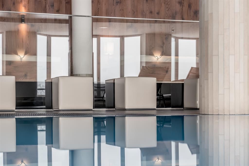 An elegant interior with modern seating and large windows. The room is reflected in the water of a pool.