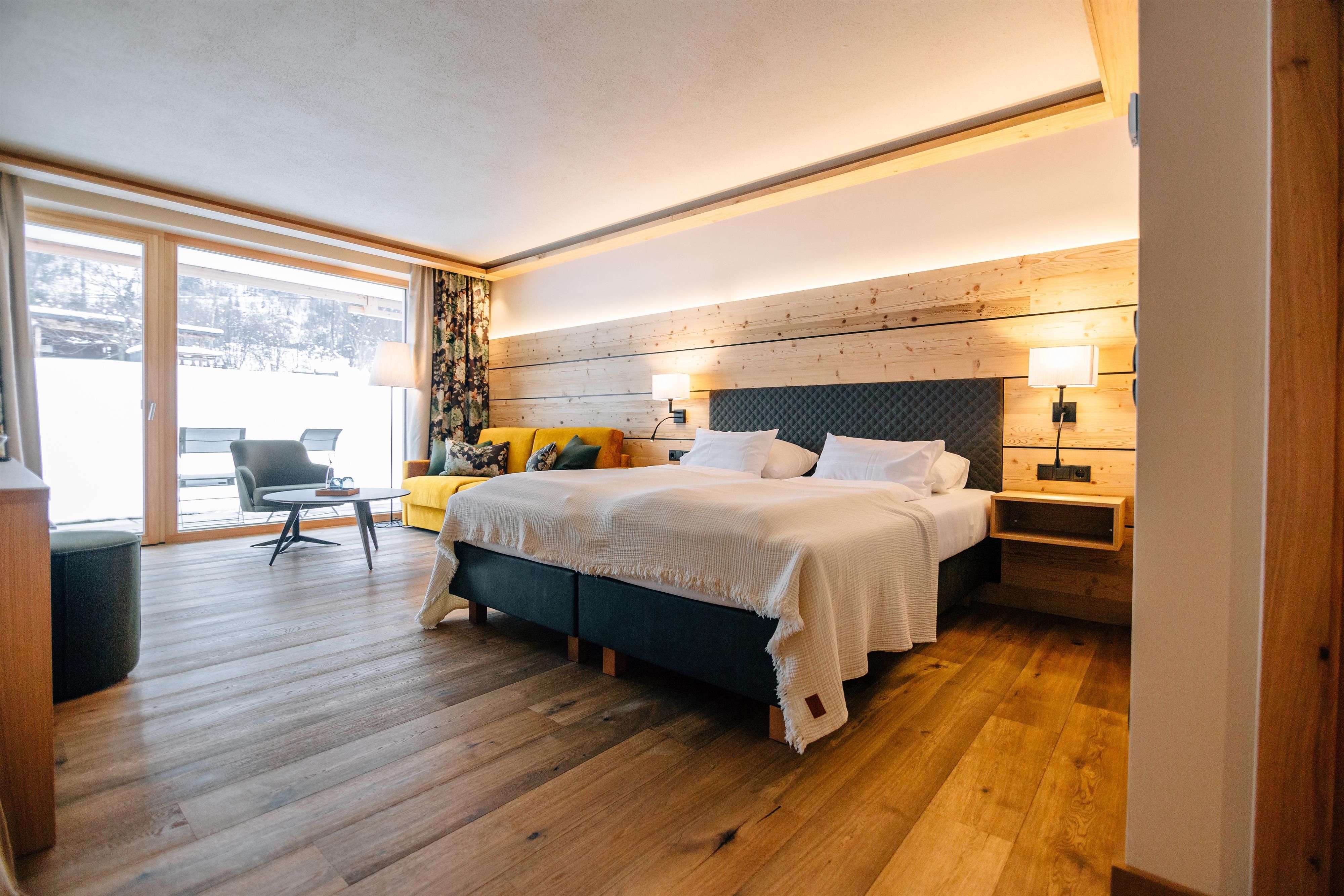 A modern hotel room with wooden ceilings and large windows. There is a comfortable bed and a cozy seating area.