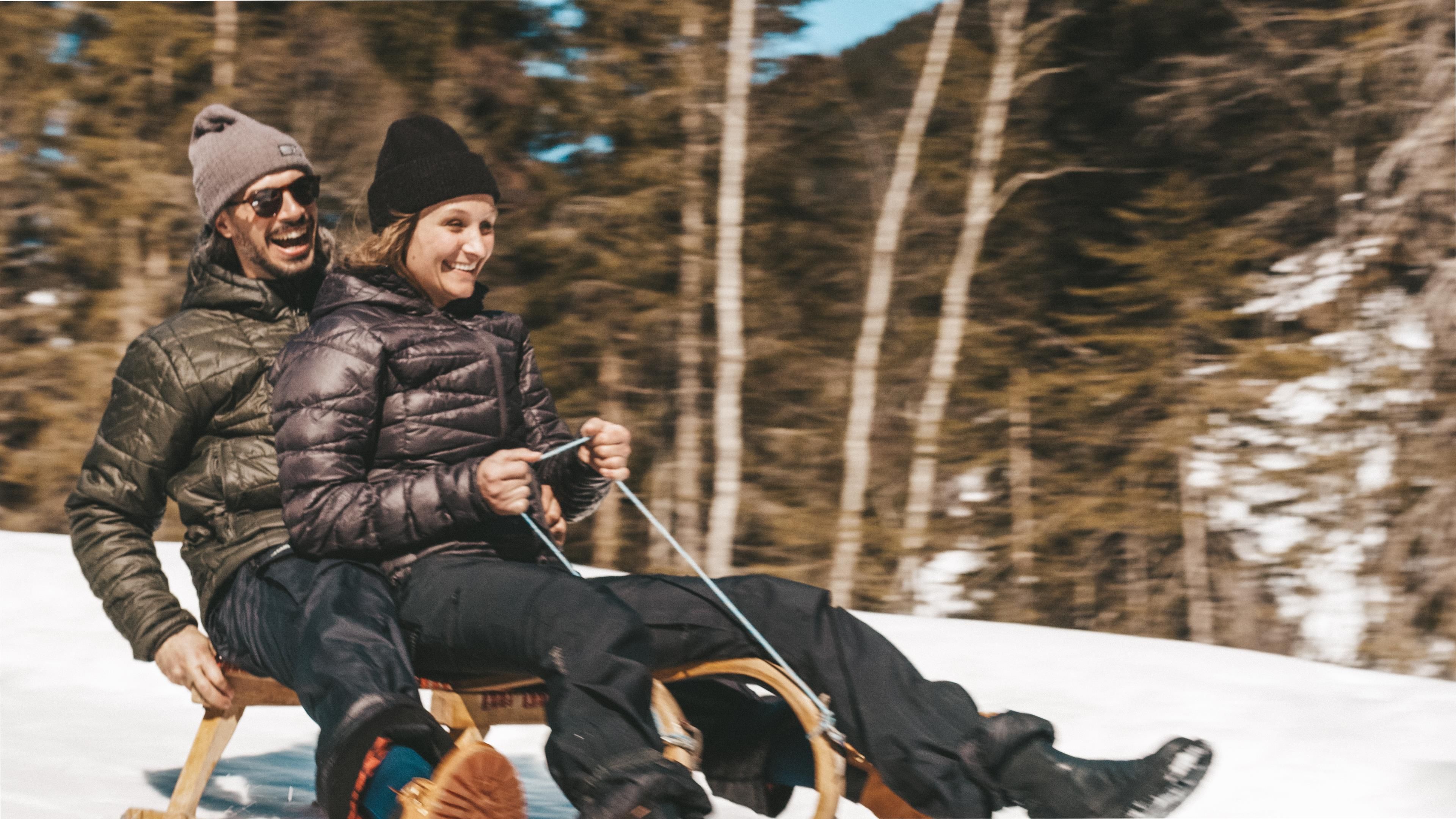 A cheerful couple rides together on a sled in the snow. The surroundings are surrounded by trees and it is a sunny day.