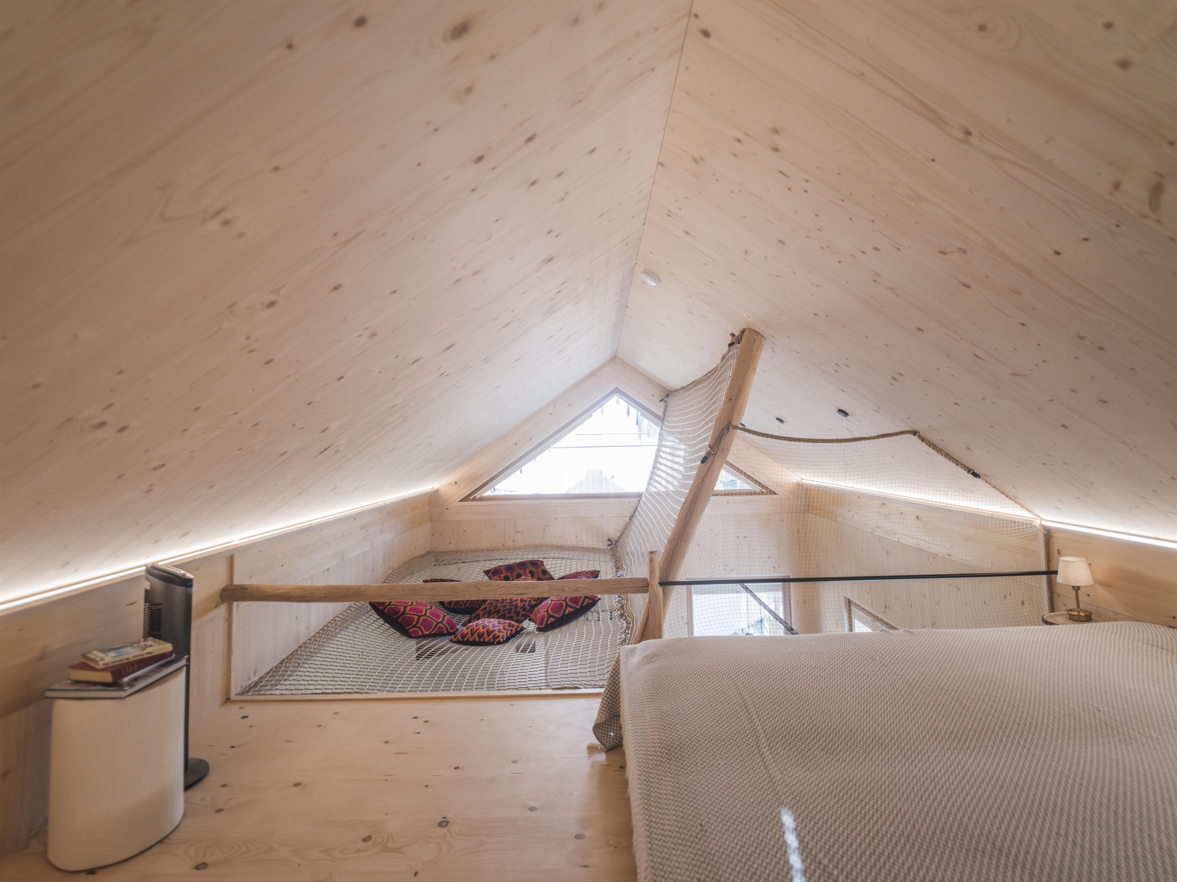 A modern, bright attic room with wooden paneling and large windows. A cozy lounging area is designed above a sleeping area.