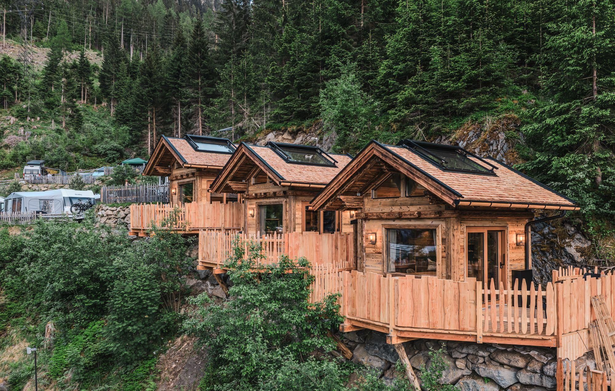 Three cozy wooden cabins surrounded by trees and rocks. They are situated on an elevated terrace with a beautiful view of nature.
