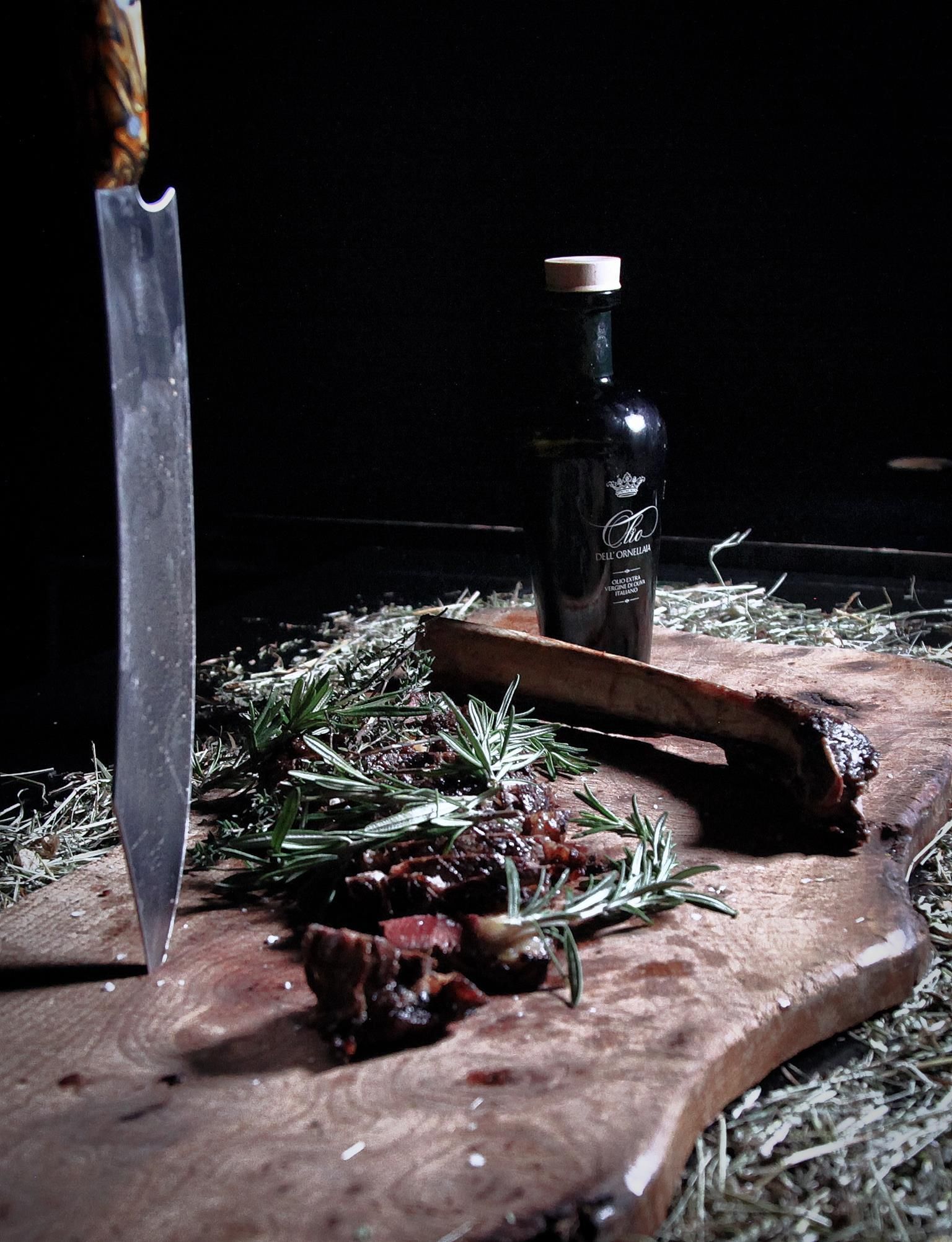 A wooden board with pieces of meat and fresh herbs lies on a dark surface. Next to the board stands a bottle of oil and a large knife.