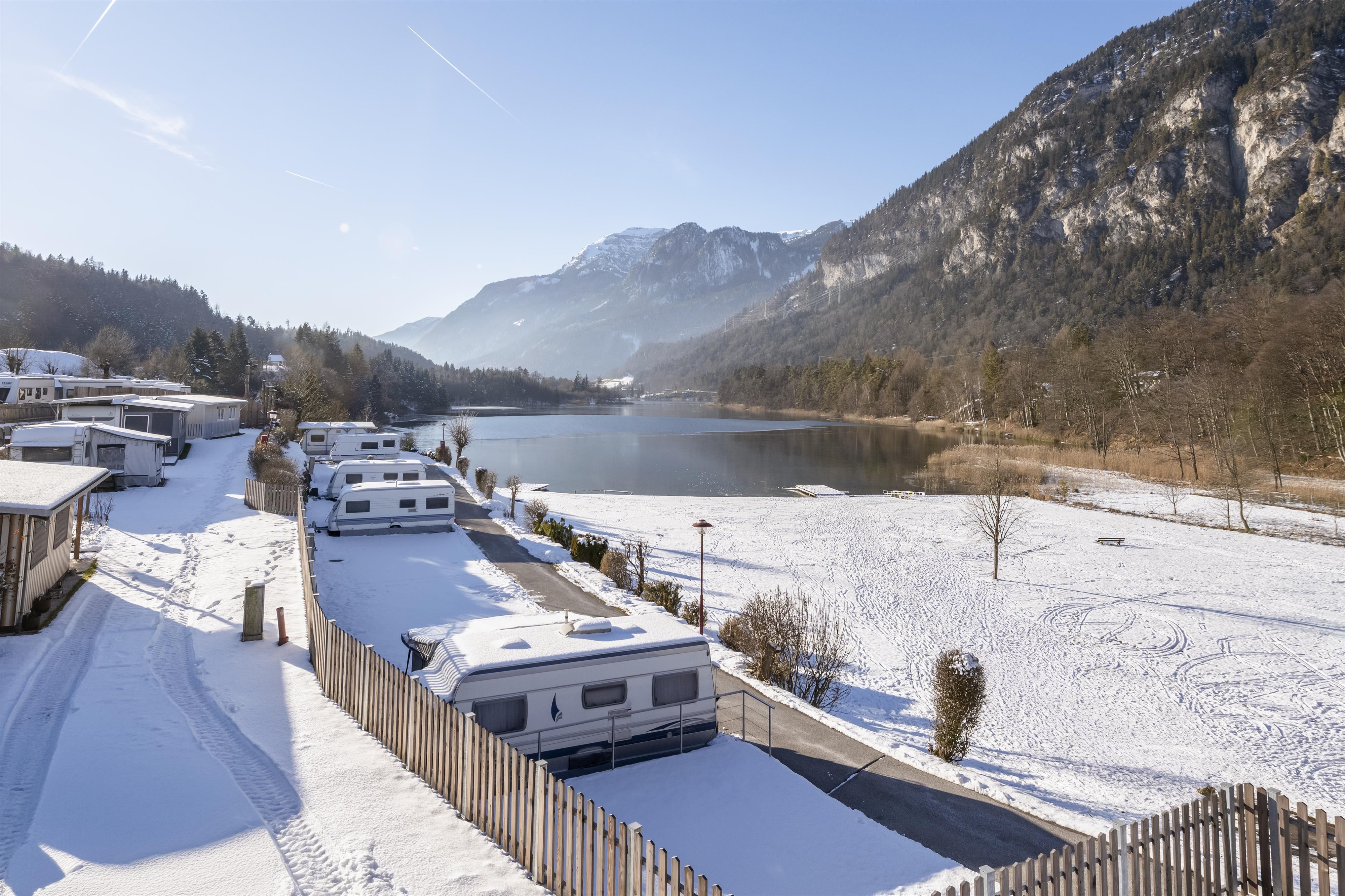 A winter landscape with snow-covered hills and a silent lake. Campervans are parked along a path, surrounded by trees.