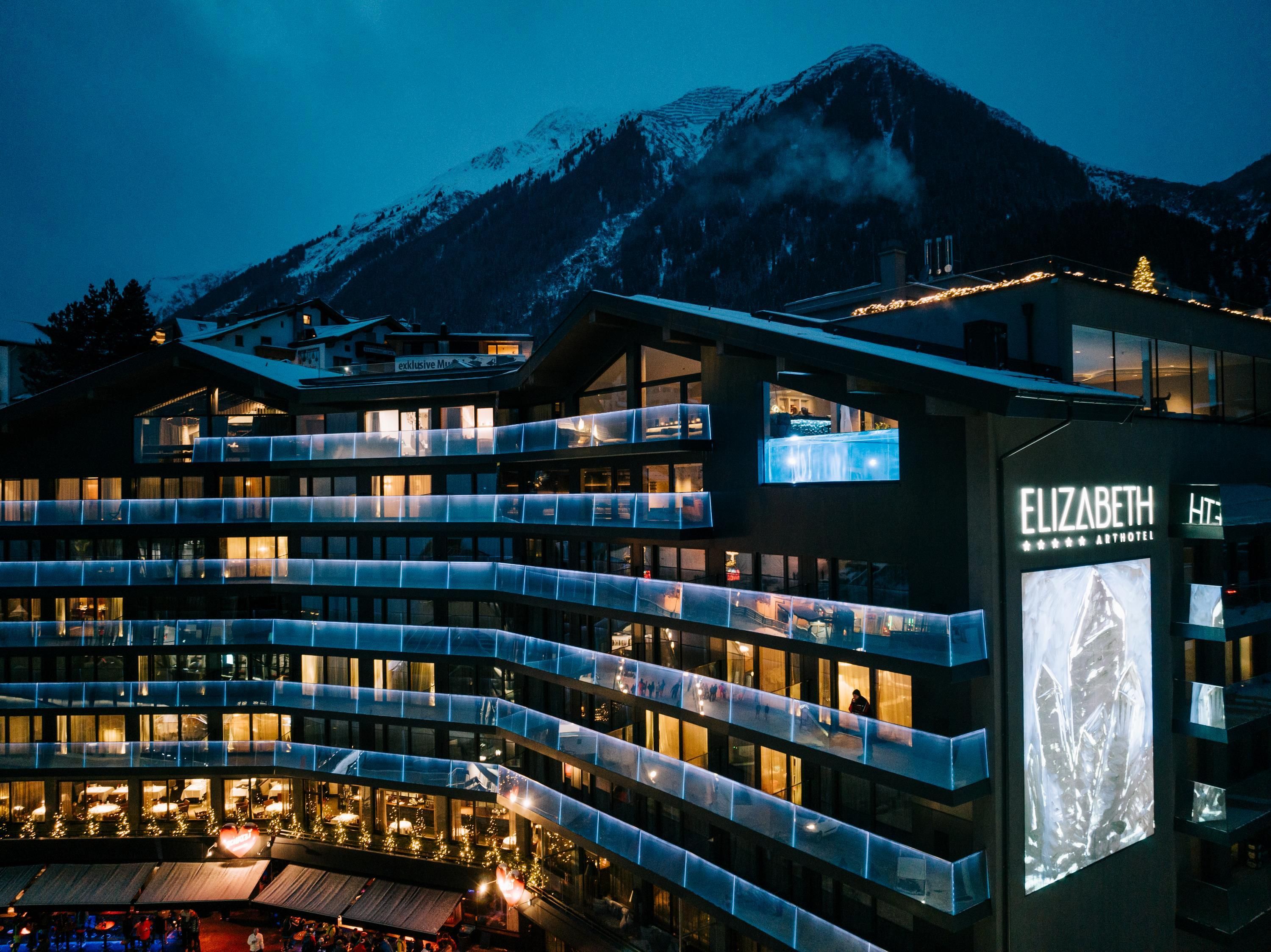 A modern hotel with illuminated balconies, surrounded by majestic mountains. The evening sky adds a calm atmosphere to the scene.