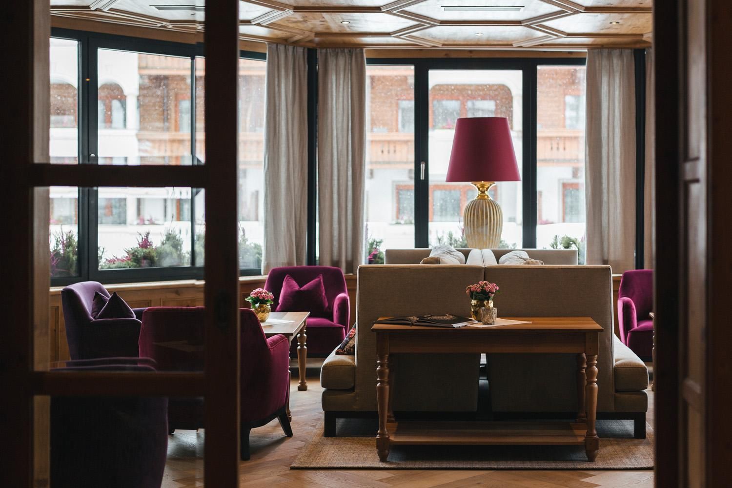 An elegant living room with comfortable purple armchairs and a large window that lets in natural light. In the center, there is a wooden table with decorative flowers.