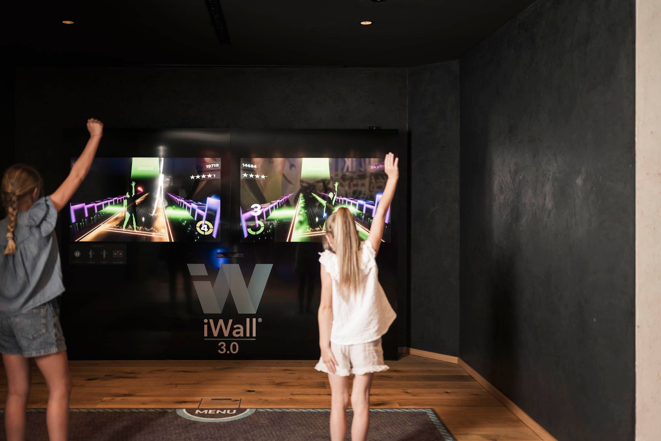 Two girls are playing an interactive game on a large screen. They are standing in front of the wall with colorful graphics and have raised their arms.