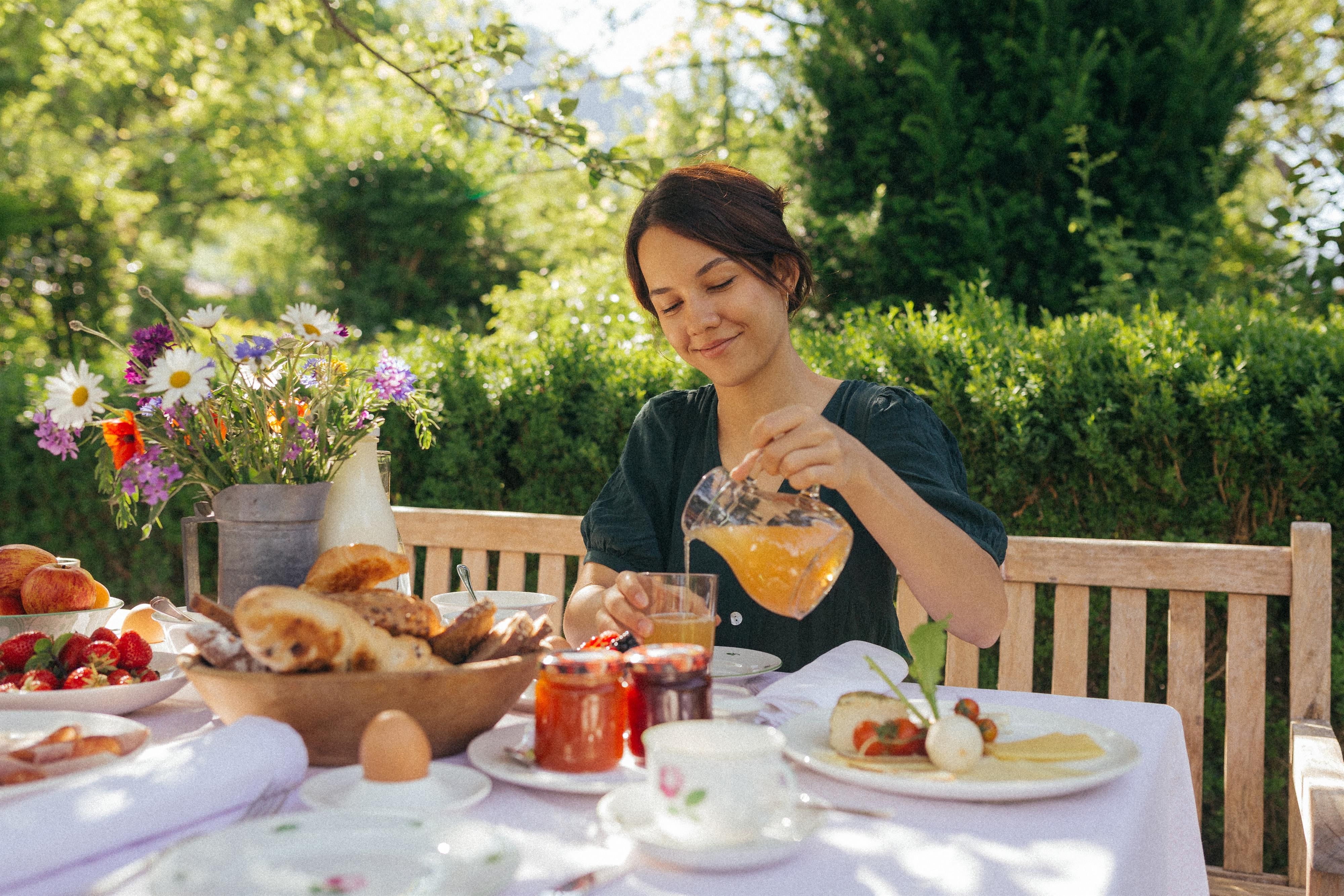 A woman is sitting at a table outdoors, pouring juice into a glass. The table is decorated with various breakfast treats and flowers.