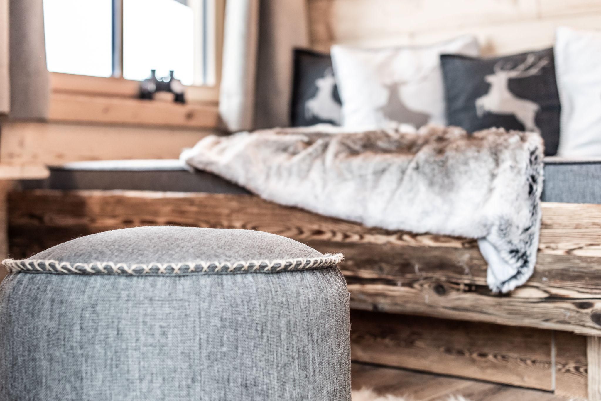 A cozy room with a wooden bed and decorative pillows. A gray stool is in the foreground, while a blanket is lying on the bed.
