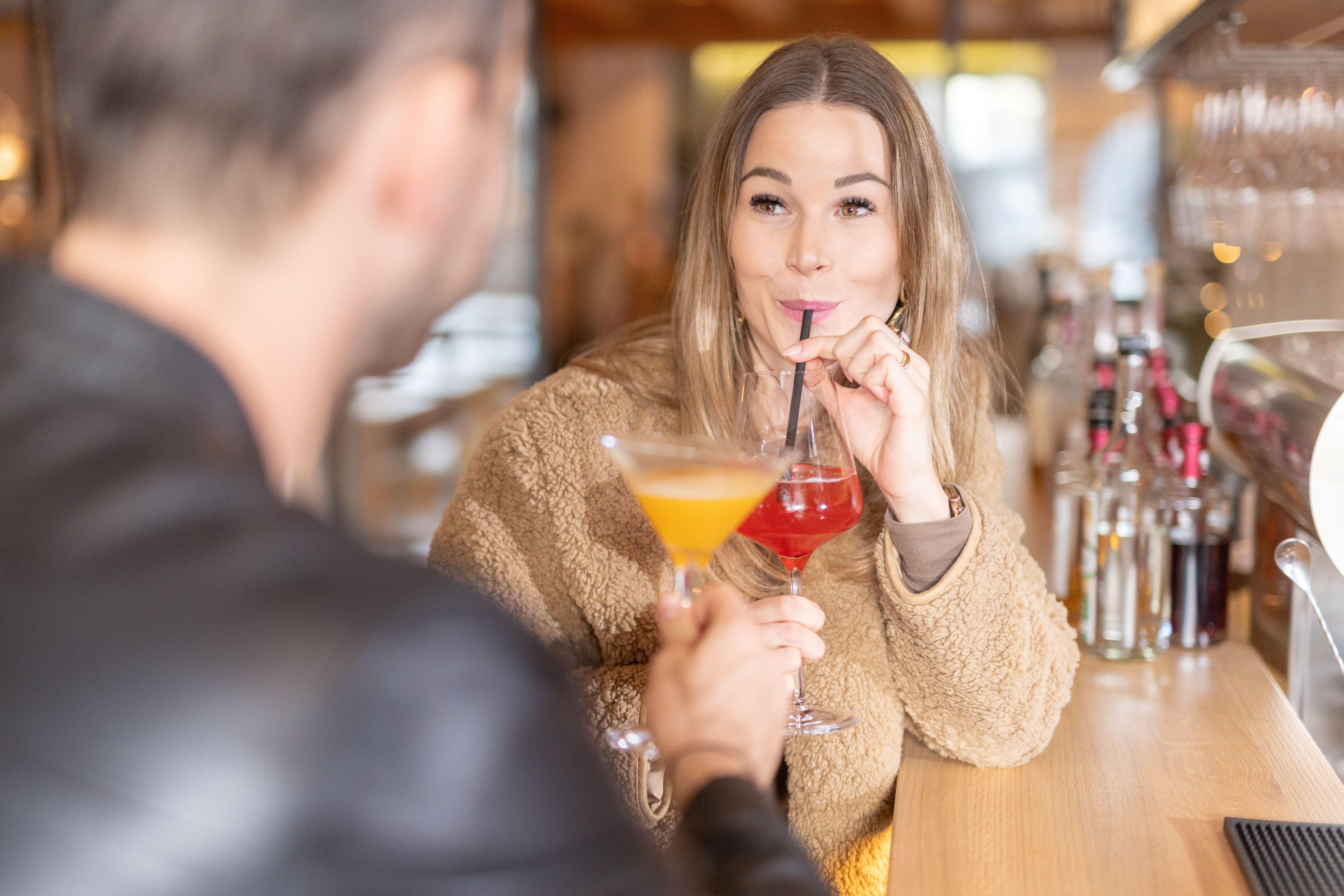 A woman enjoys a cocktail at the bar and smiles. In the background, more drinks are visible.