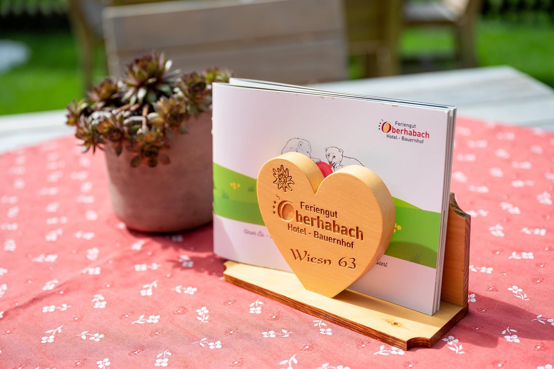 A table with a heart-shaped wooden decoration and a brochure about the hotel "Berthia-Bach". In the background, there is a pot with a succulent.