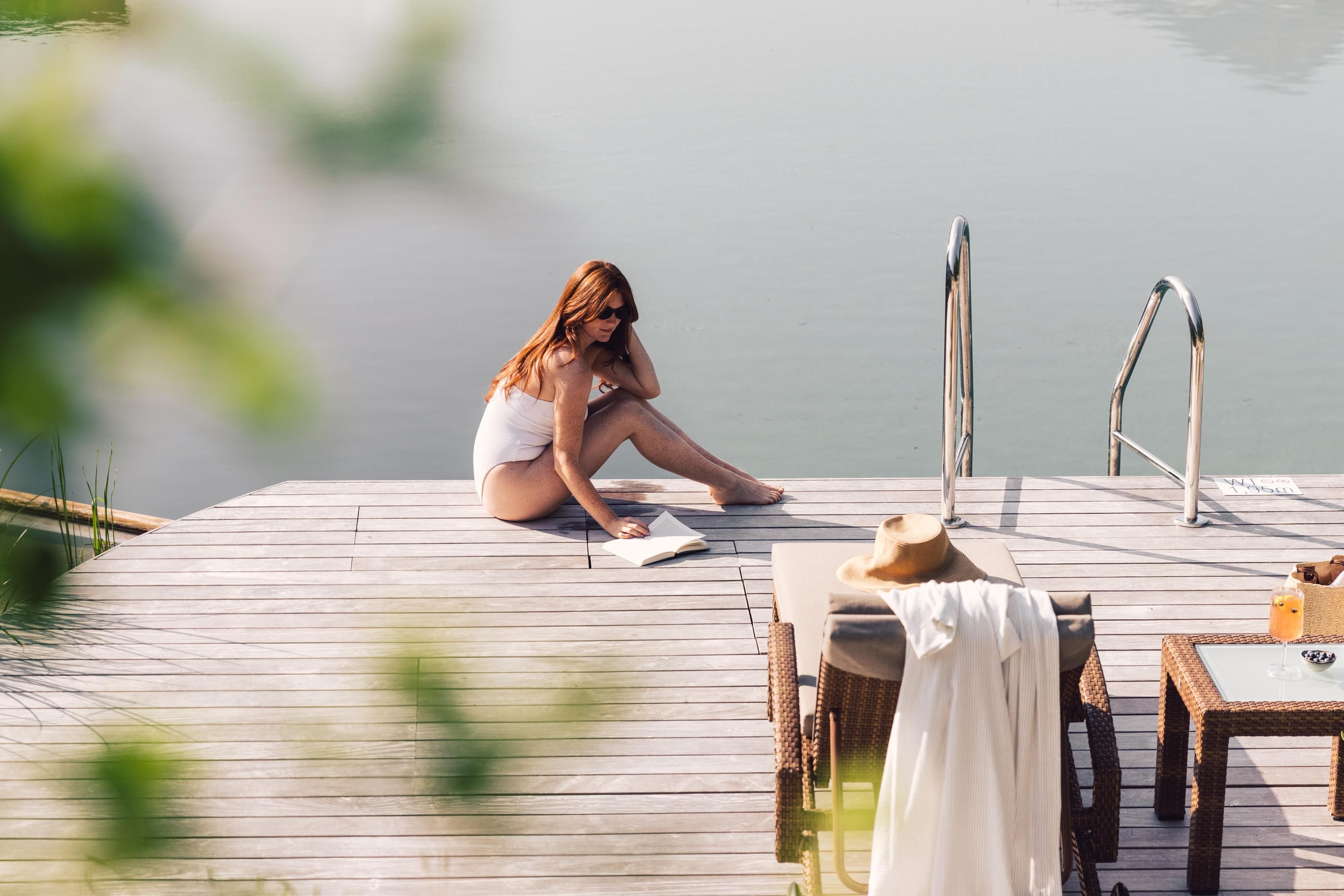 A woman is sitting on a wooden dock by the water, reading a book. Next to her are a sturdy sun umbrella and a towel.