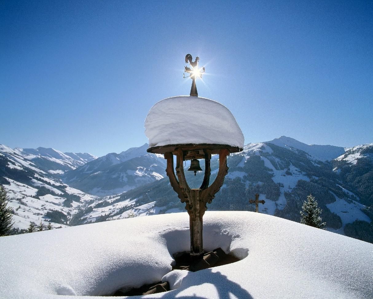 A winter landscape with snow-covered mountains and a bright blue sky. In the foreground stands a bell in a wooden frame, covered with snow.