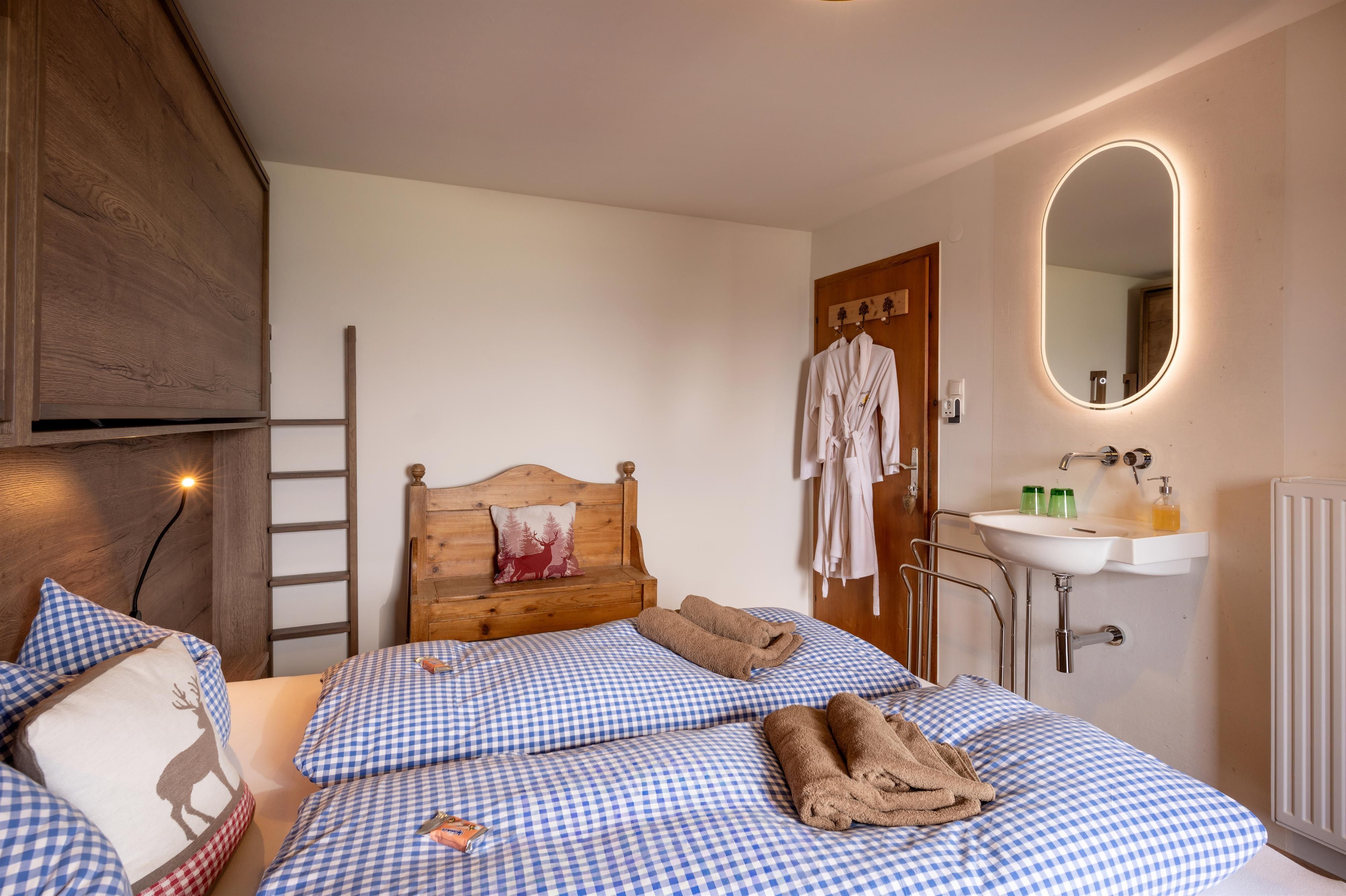 A cozy bedroom with a double bed and blue-white checks. There is an armchair, a sink, and a large mirror on the wall.