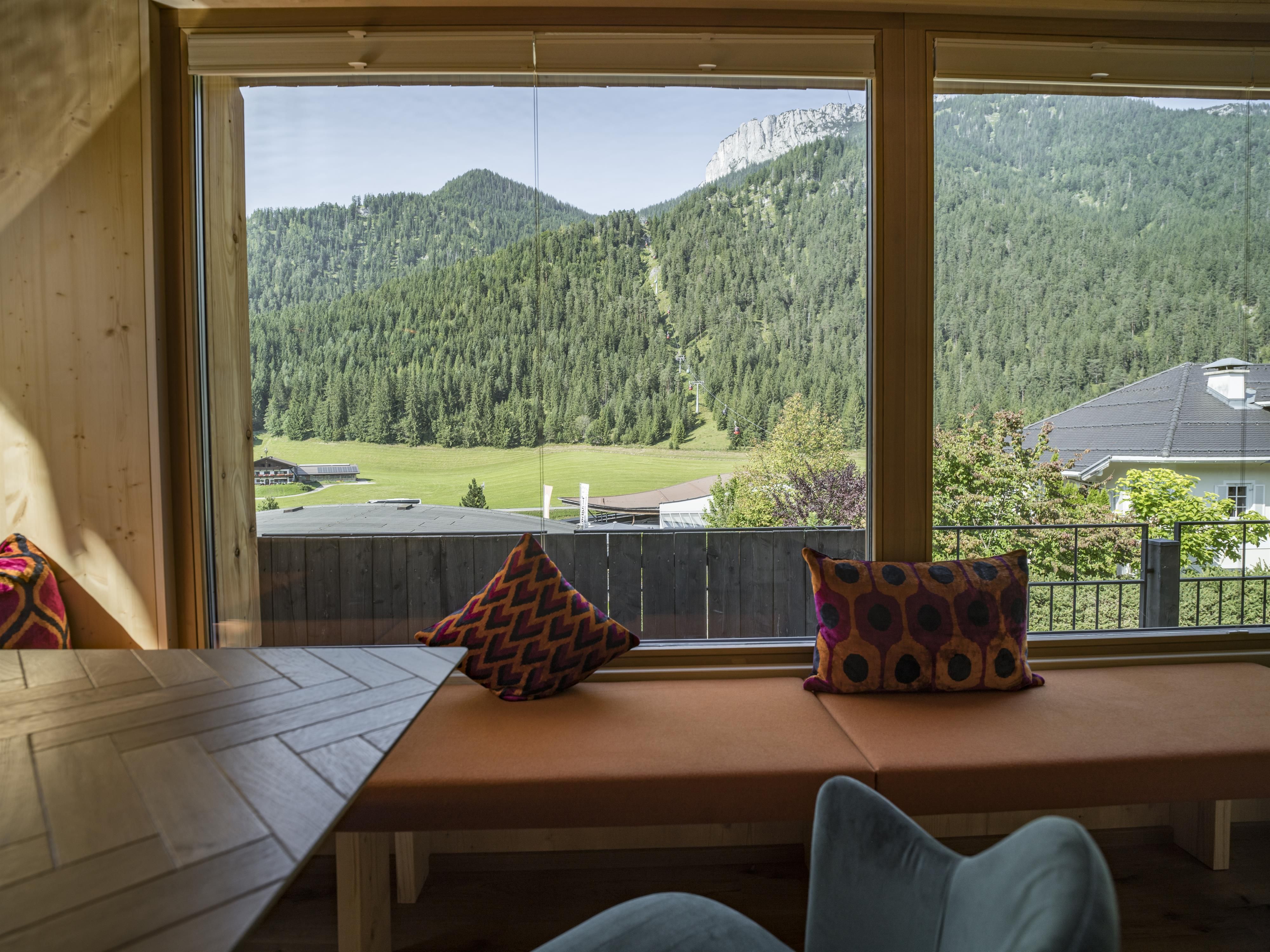 A cozy interior with a large window front that offers a view of green mountains and meadows. Colorful cushions are placed on the windowsill.