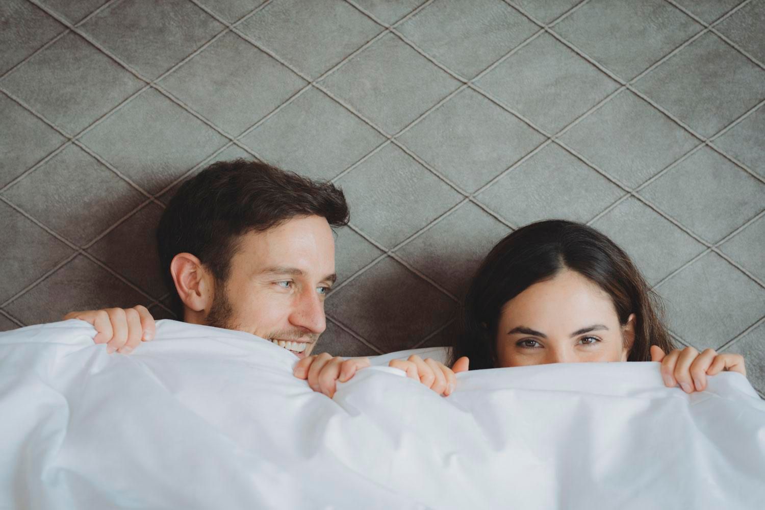 A happy couple smiles as they hide under a blanket. They display a loving and playful mood.