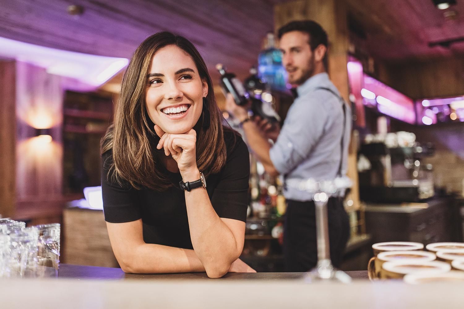 A smiling woman at the bar with a bartender in the background. The surroundings have a modern, cozy atmosphere with warm colors.