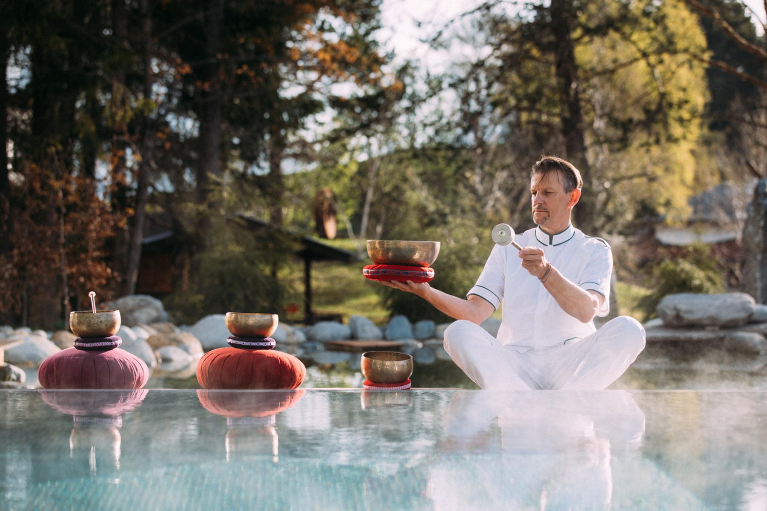 A man sits in the snow lotus on a glass surface and plays a sound bowl. In the background, trees and a calm environment can be seen.