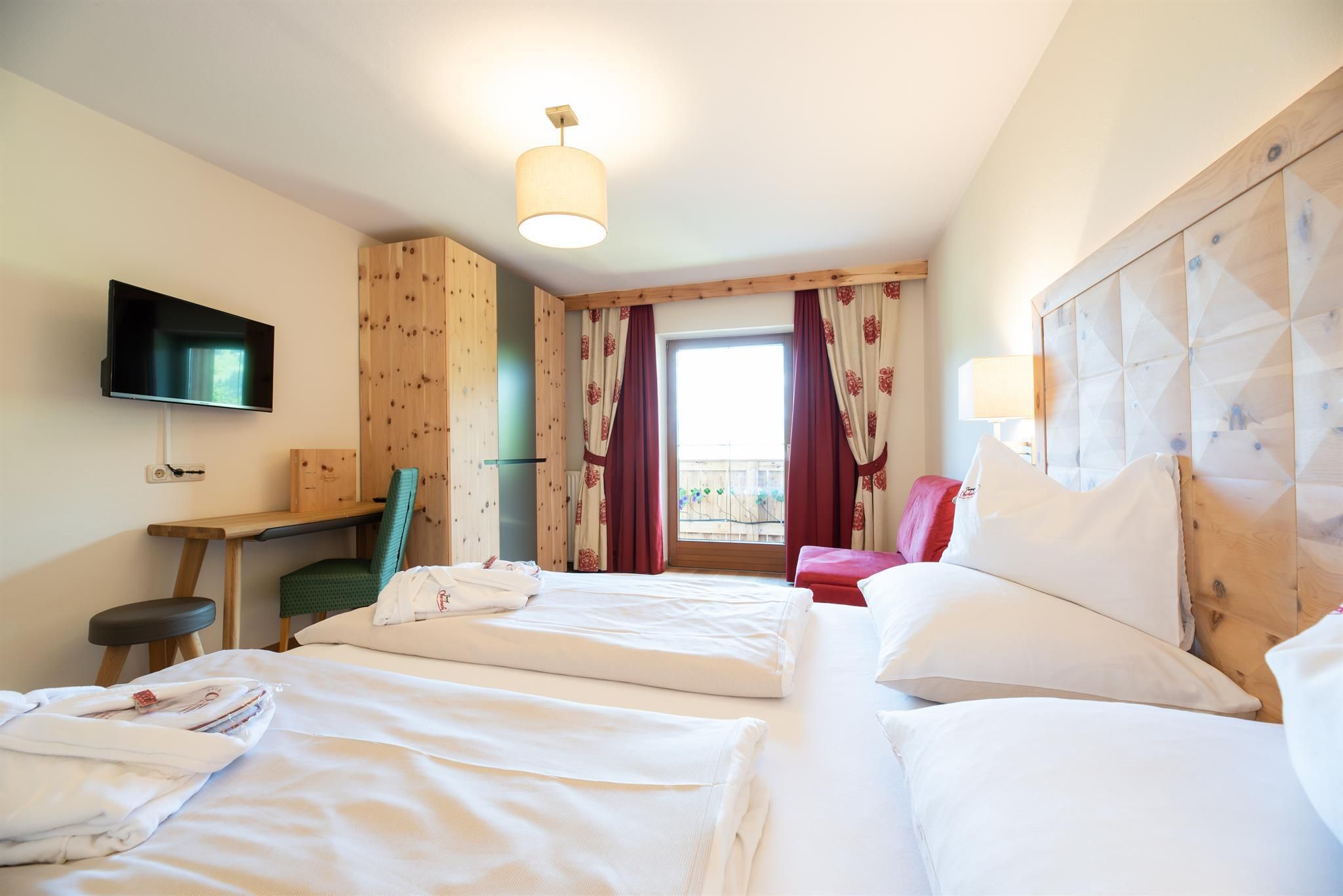 A cozy hotel room with two beds and a desk. There is a television and a view of the balcony.