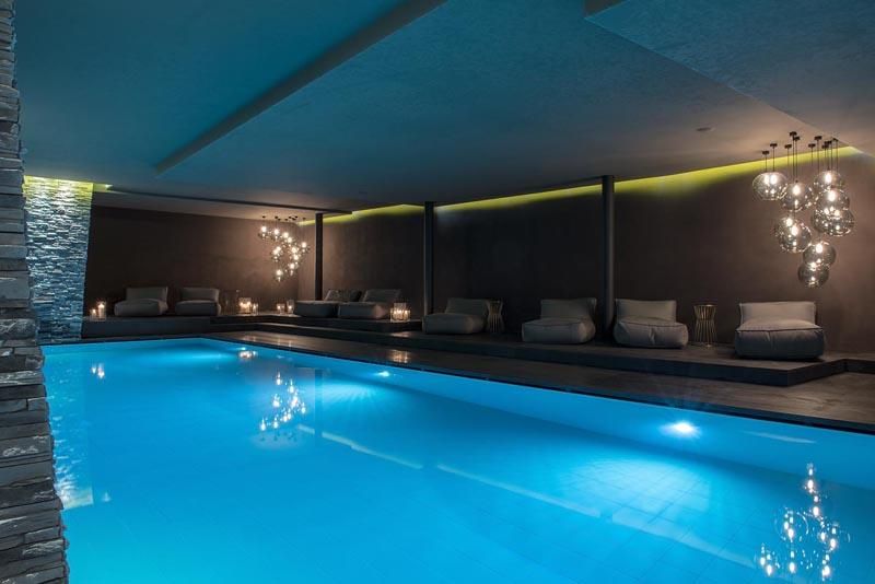 An elegant indoor pool area with soft, blue lighting. Cozy loungers and stylish lighting create a relaxing atmosphere.