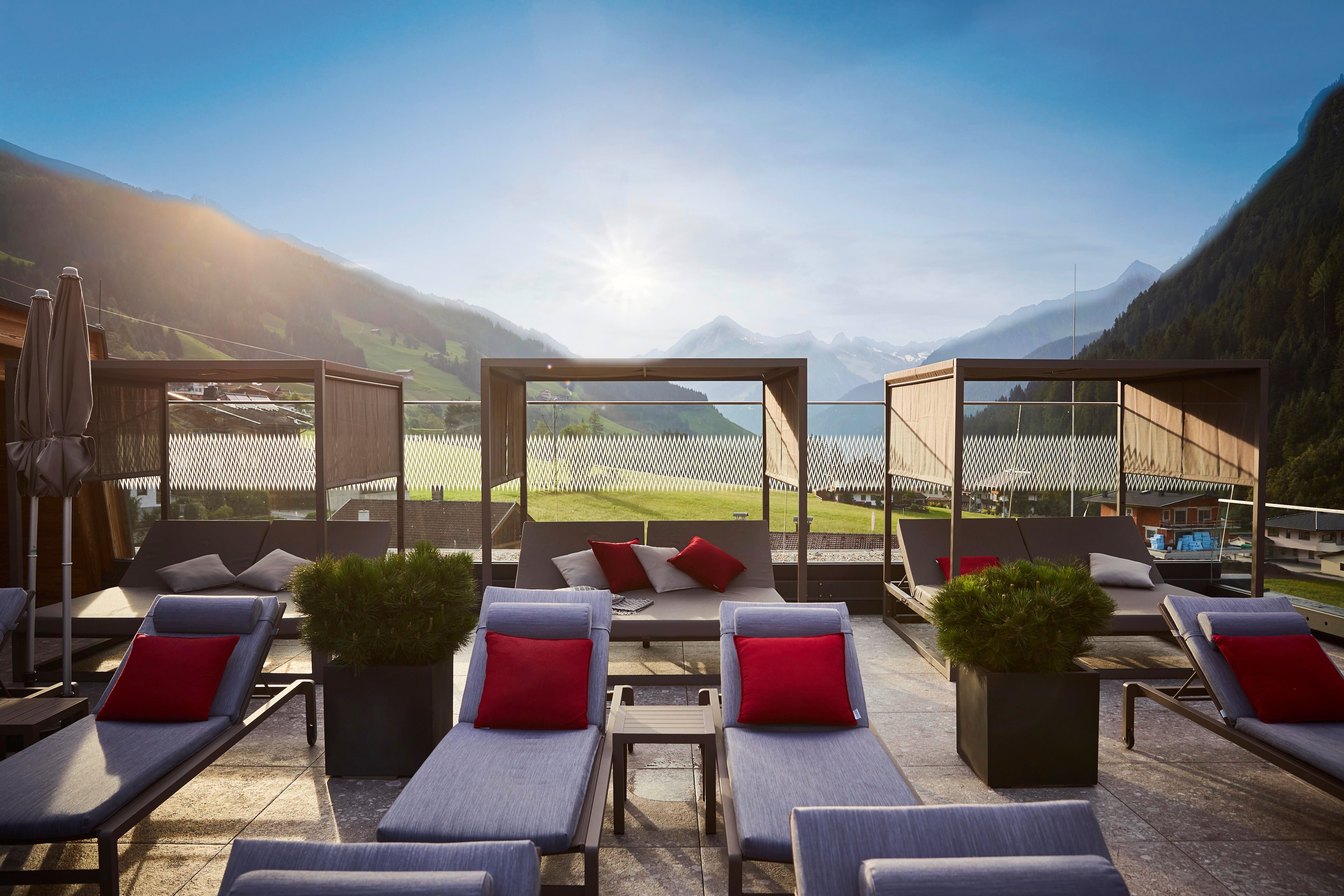 A stylish rooftop terrace with loungers and cabanas. In the background, green mountains and a clear sky stretch out.