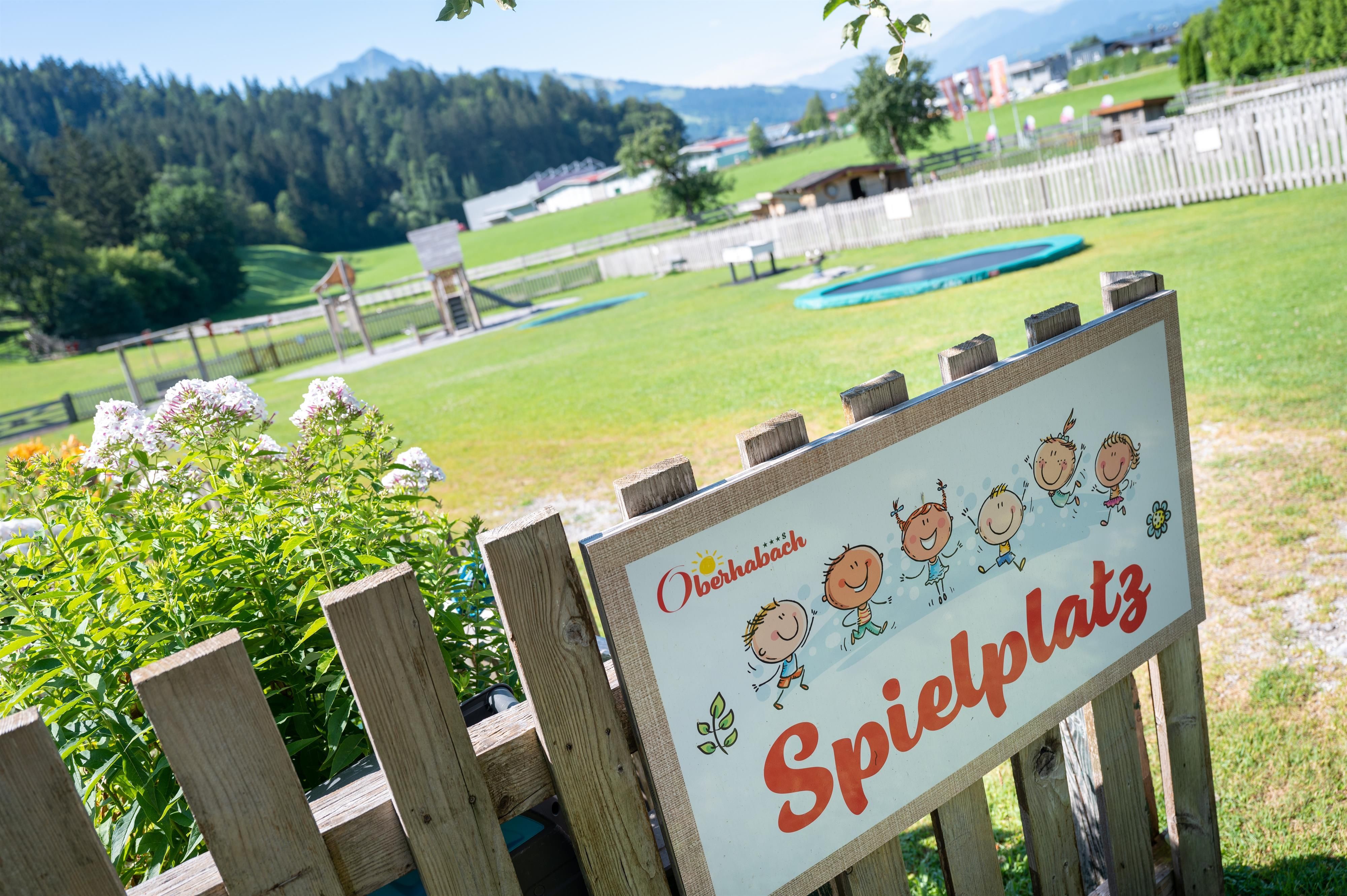 A playground with various play equipment and a large lawn. In the background, mountains and a play field can be seen.