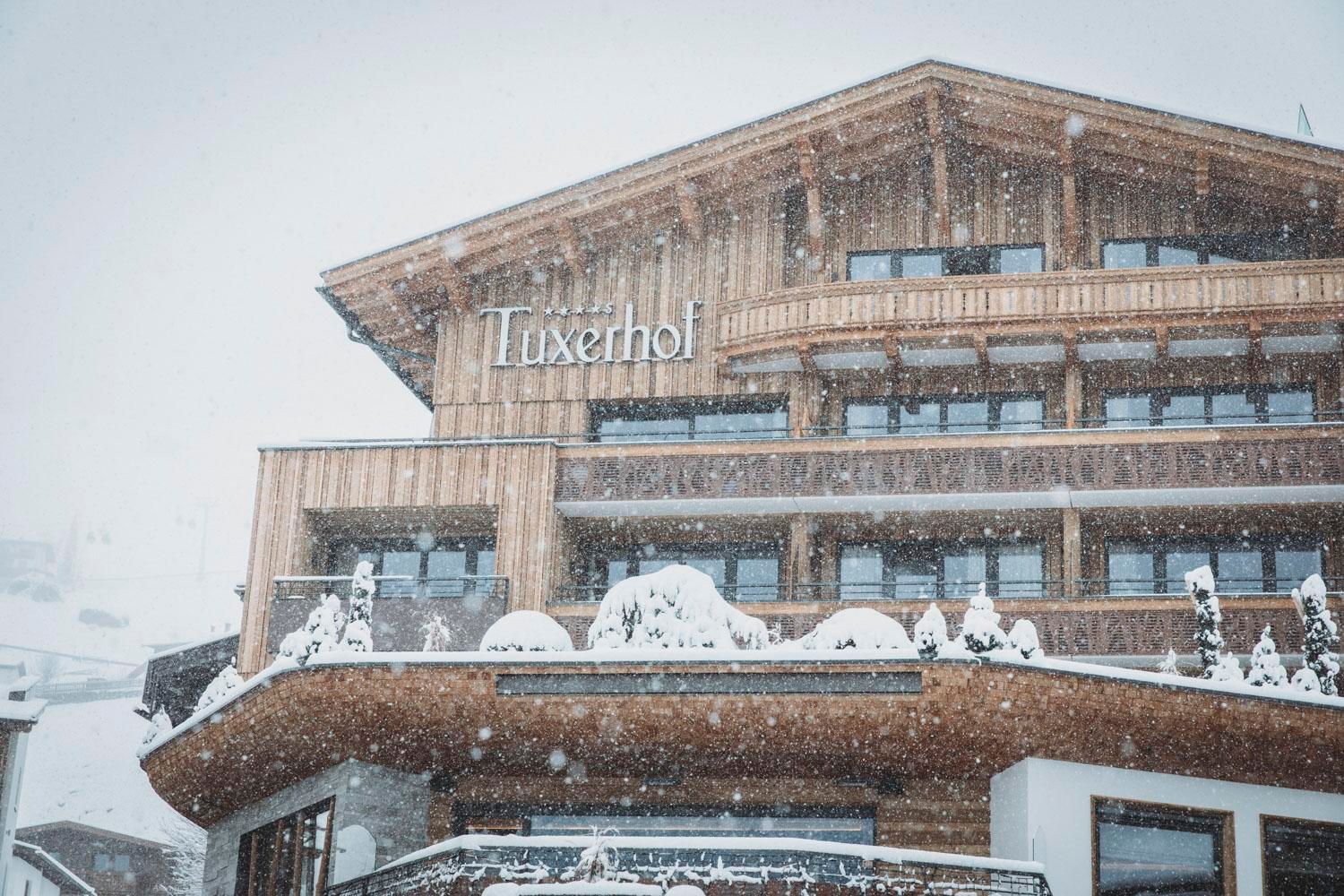 A large, modern building with a wooden facade, exposed to heavy snowfall. The name "Tuxerhol" is clearly visible on the facade.