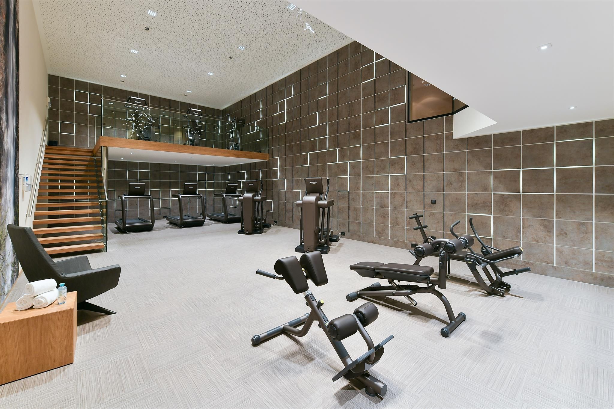 A modern gym equipped with various training machines. The walls are tiled with large gray tiles, providing a bright, clean environment.