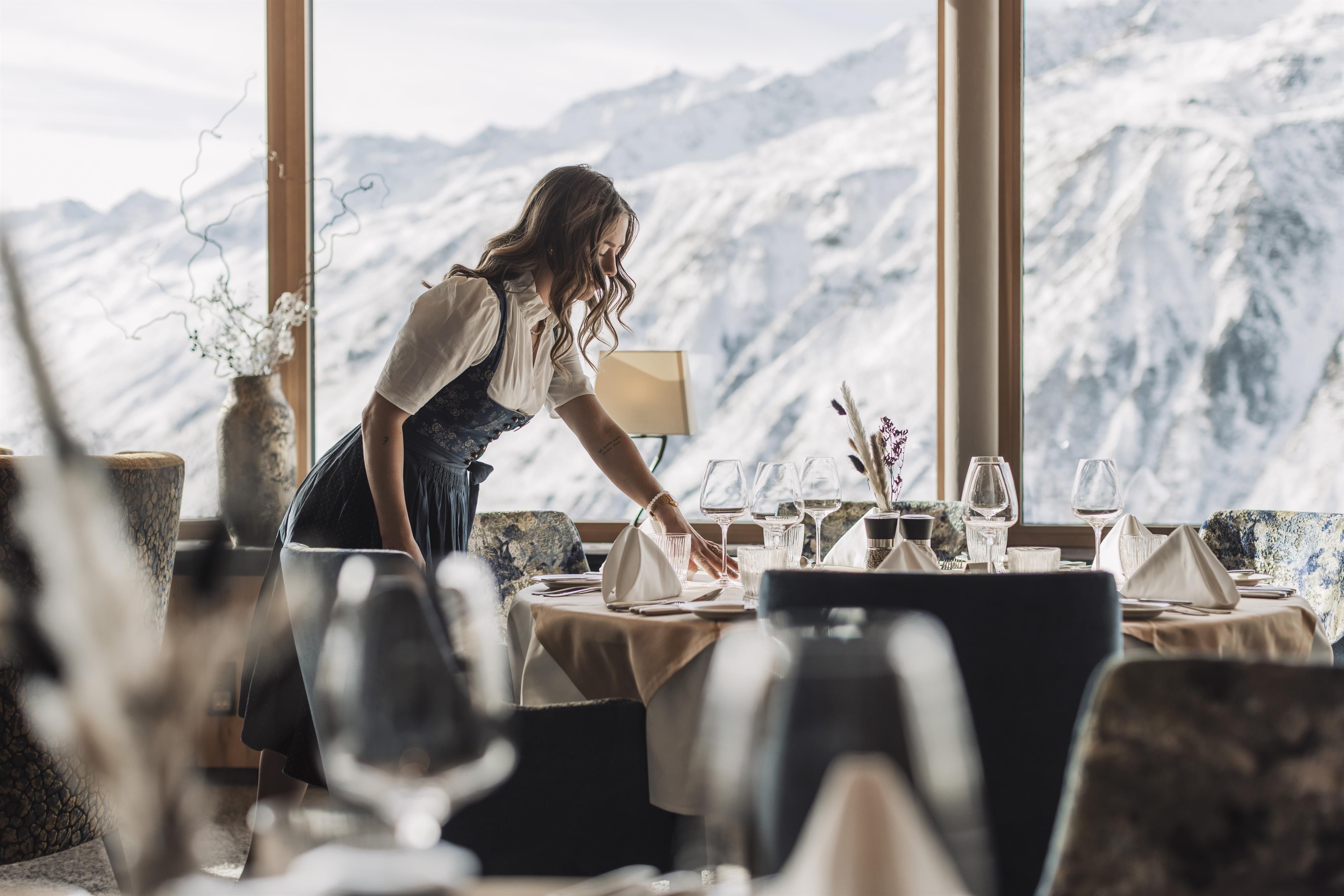 A waitress is setting a table in a restaurant with a view of snow-covered mountains. The atmosphere is cozy and inviting.