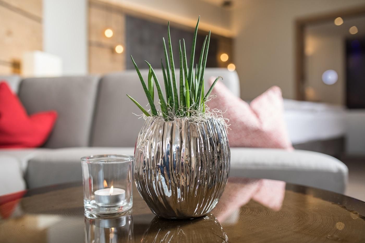 A modern living space with an elegant gray sofa and colorful cushions. On a table, there is a decorative flower pot with green plants and a candle.