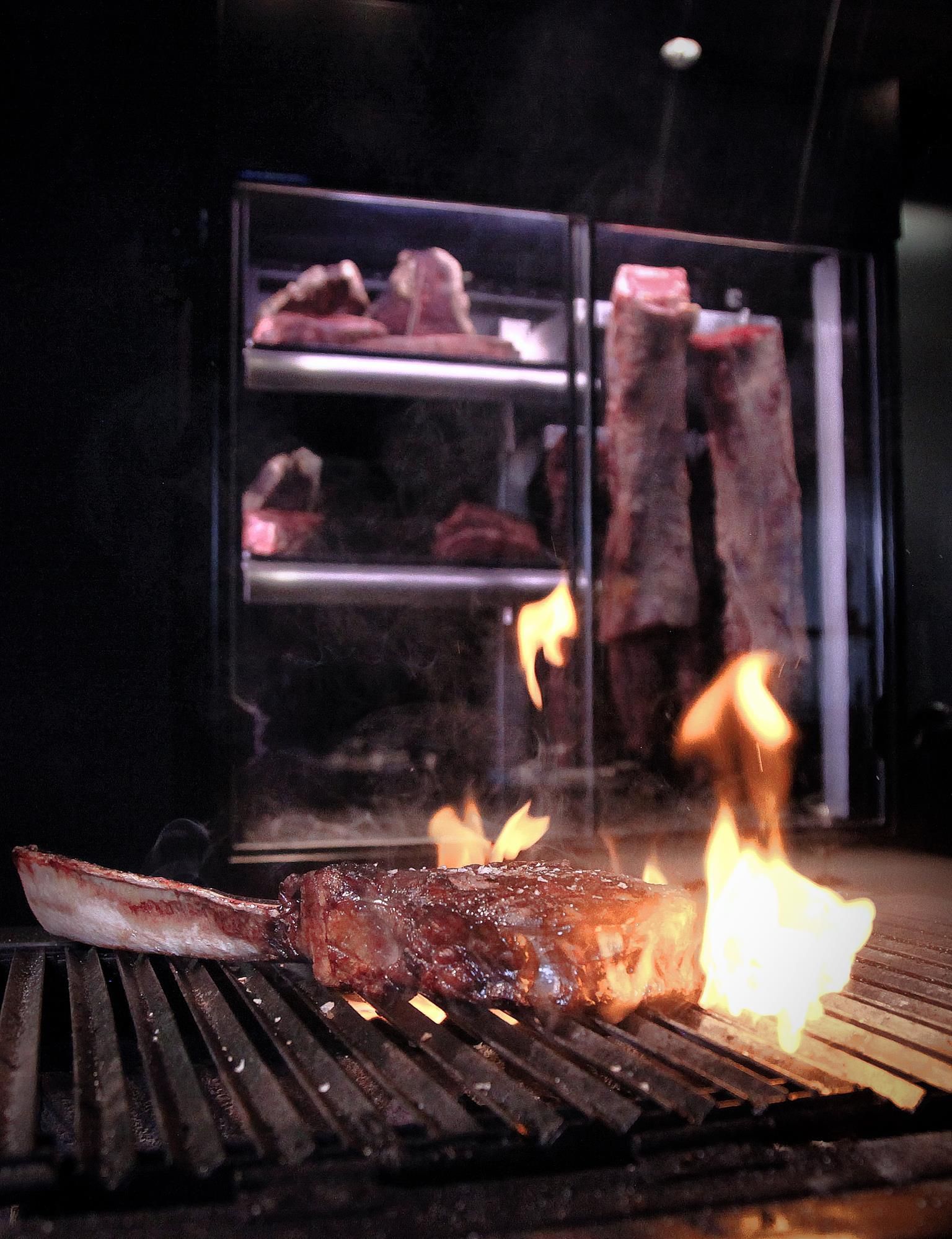 A piece of meat sizzles on a grill with flames. In the background, large pieces of meat are visible in a refrigerator.