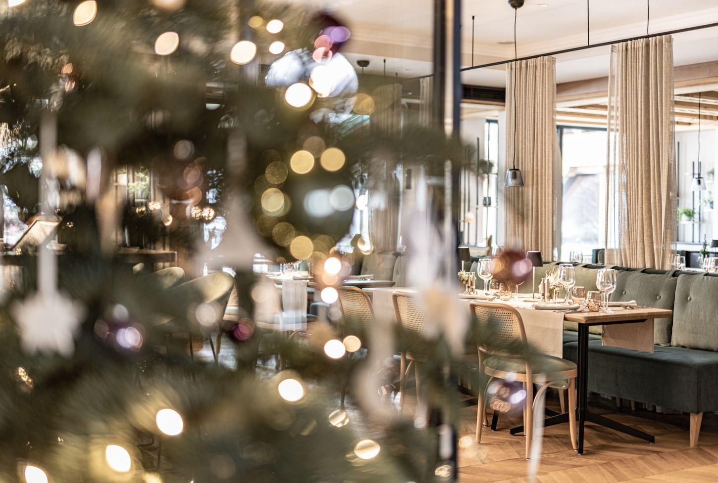 A festively decorated table in an elegant restaurant with soft lighting. In the foreground, there is a Christmas tree with lights and decorative elements.
