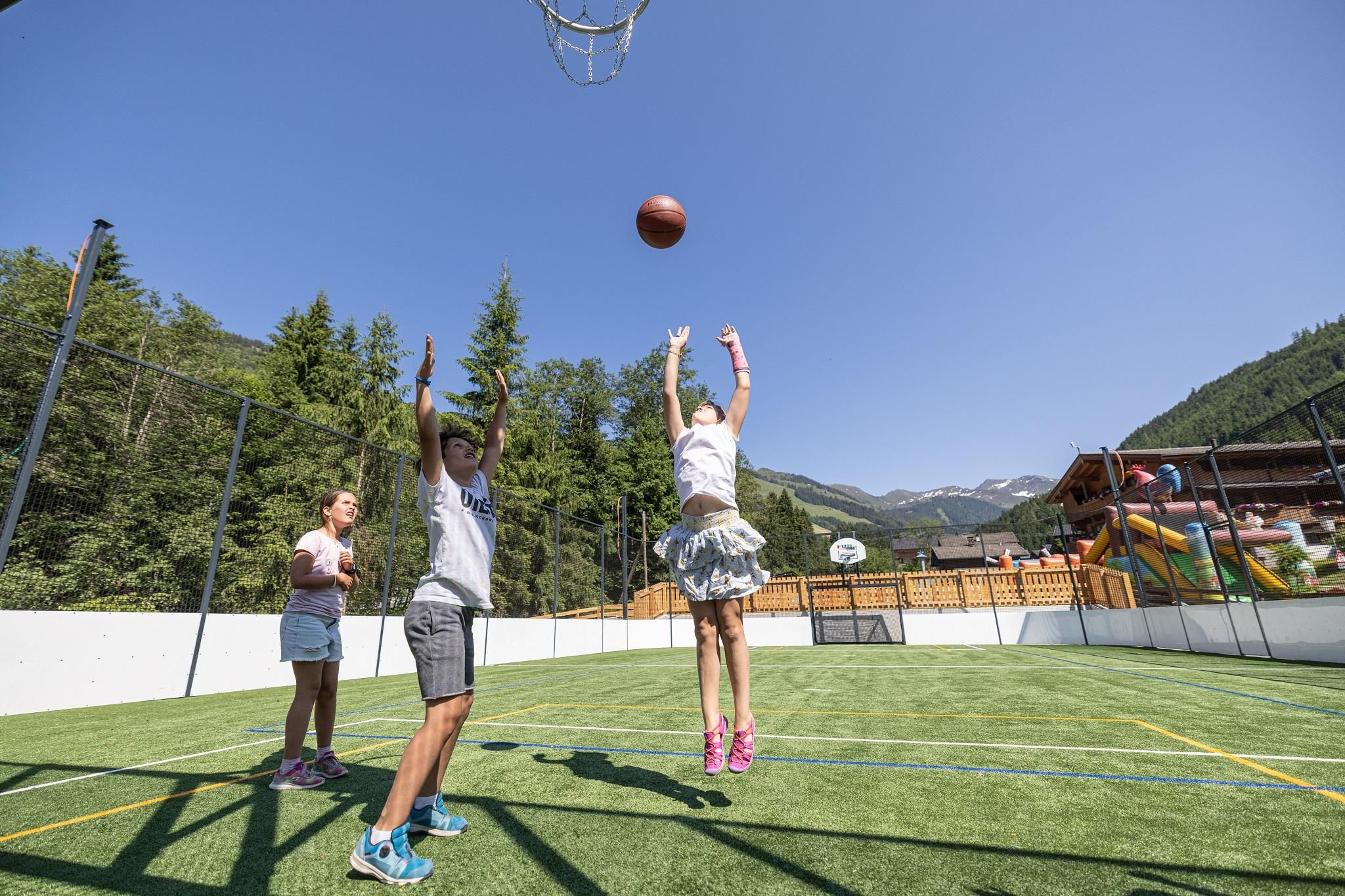 An outdoor basketball court with children playing. In the background, trees and a recreational area are visible.