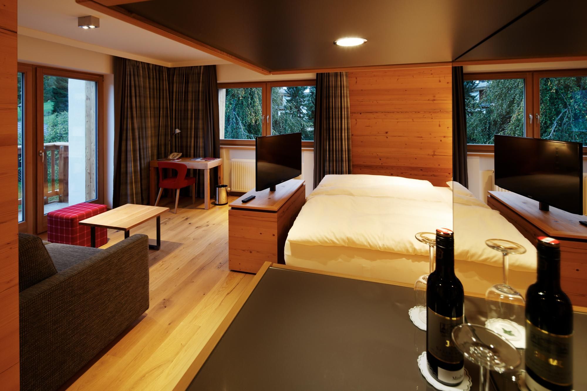 A modern hotel room with wooden furniture and large windows. The room has a cozy seating area and is brightly lit.