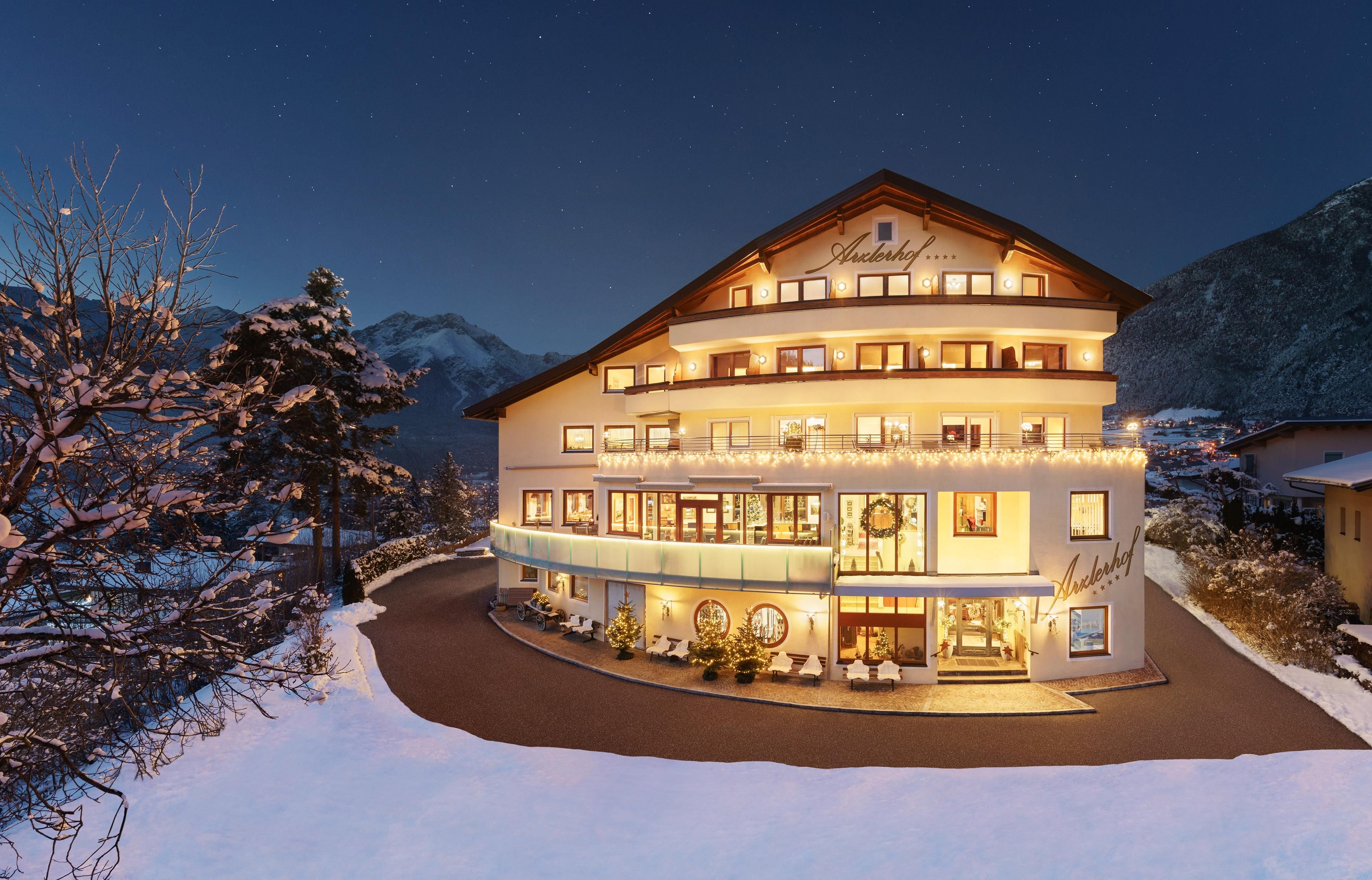 A beautiful hotel in winter with snow-covered outdoor terraces and warm light coming from the windows. The surrounding mountain landscape adds to the inviting atmosphere.