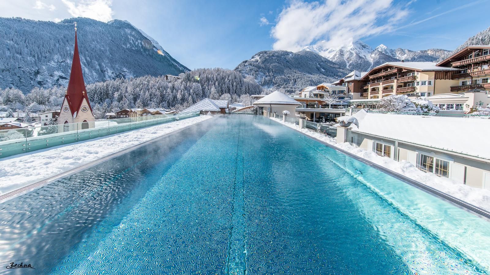 An impressive swimming pool with clear water amidst a snowy mountain landscape. Well-maintained buildings and a church complement the winter scenery.