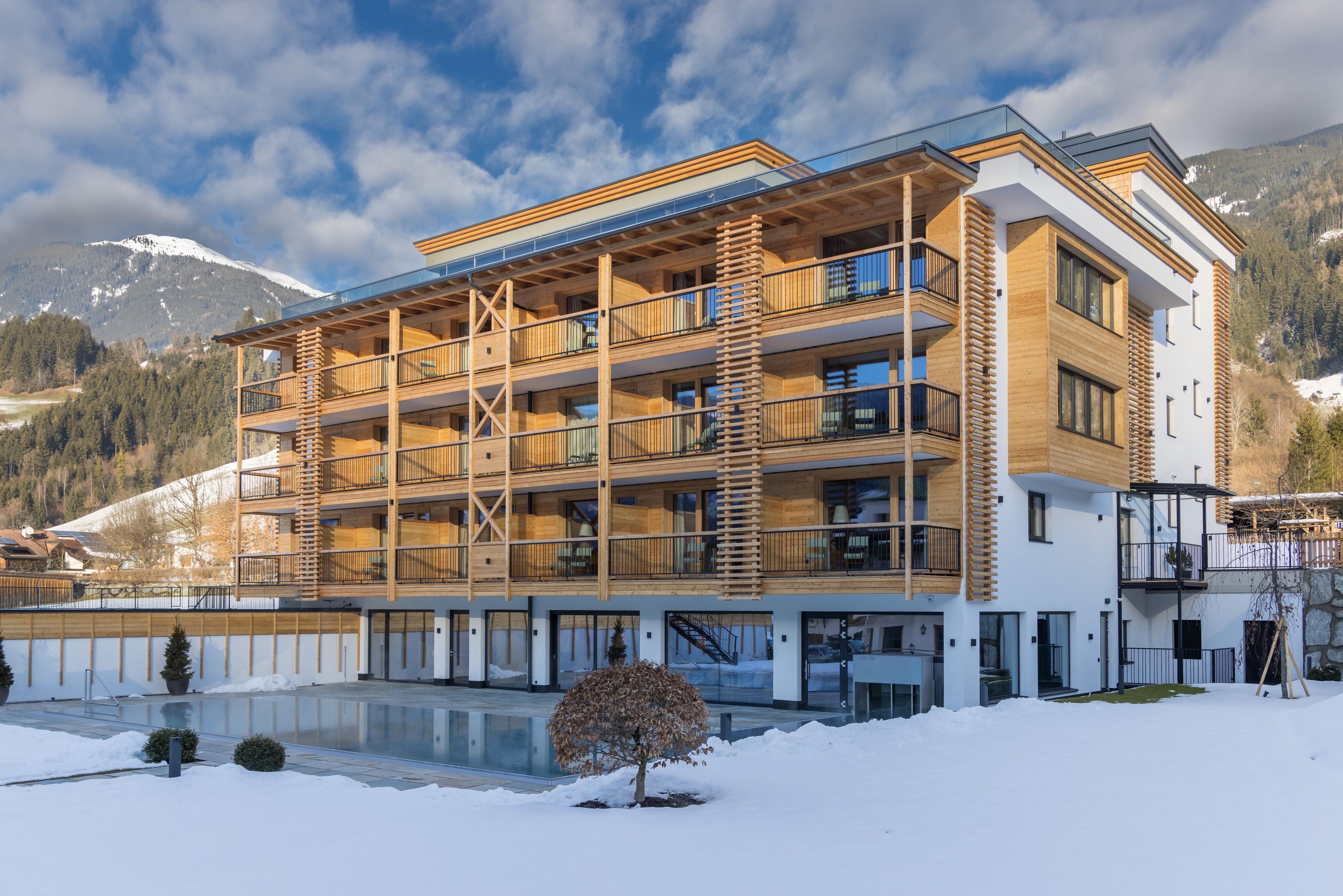 A modern hotel building with wooden elements in a snow-covered environment. In the background, mountains and a blue sky can be seen.