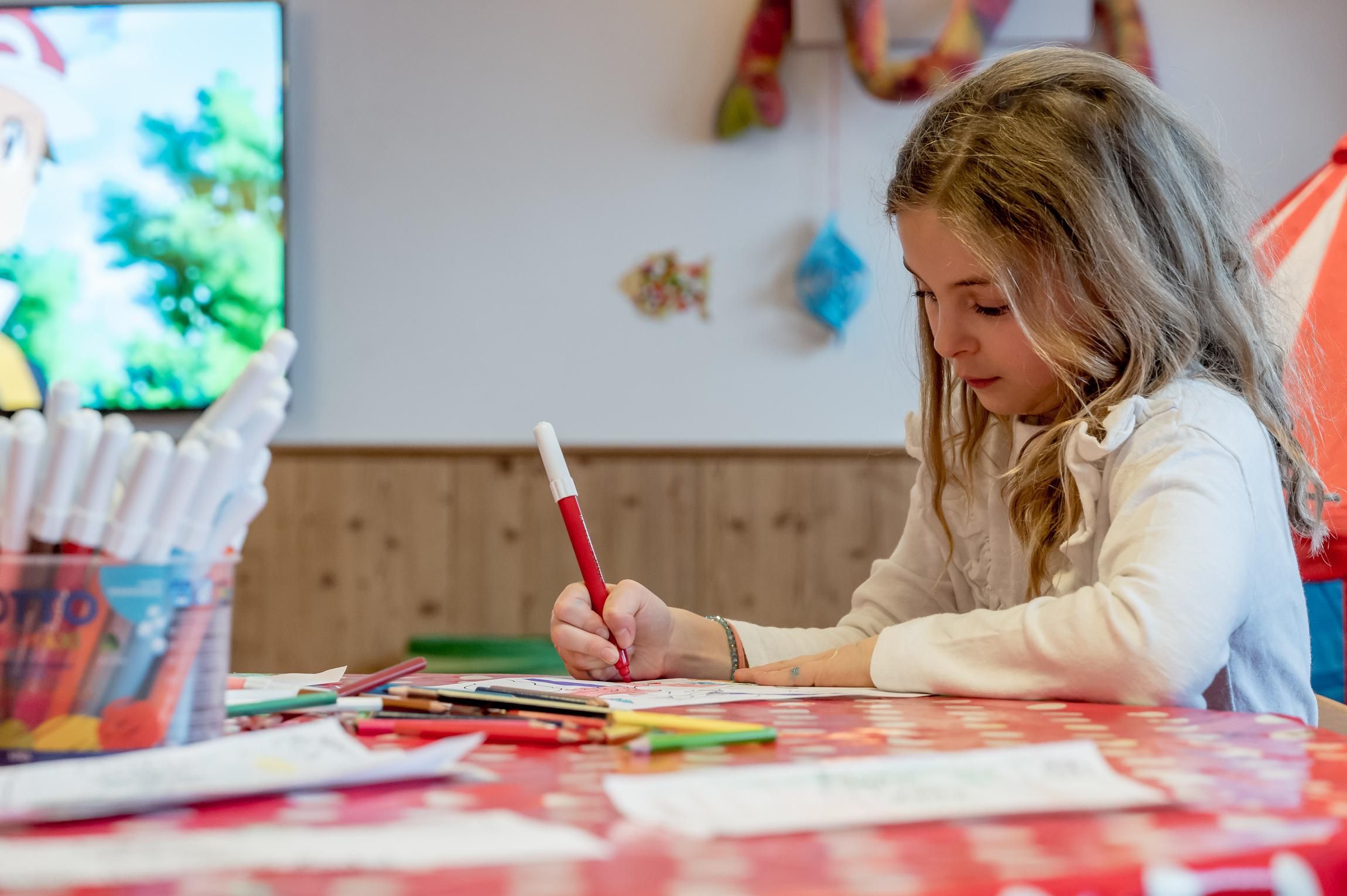 A girl is concentrating on painting at a table with colorful pencils and paper. A television is visible in the background.