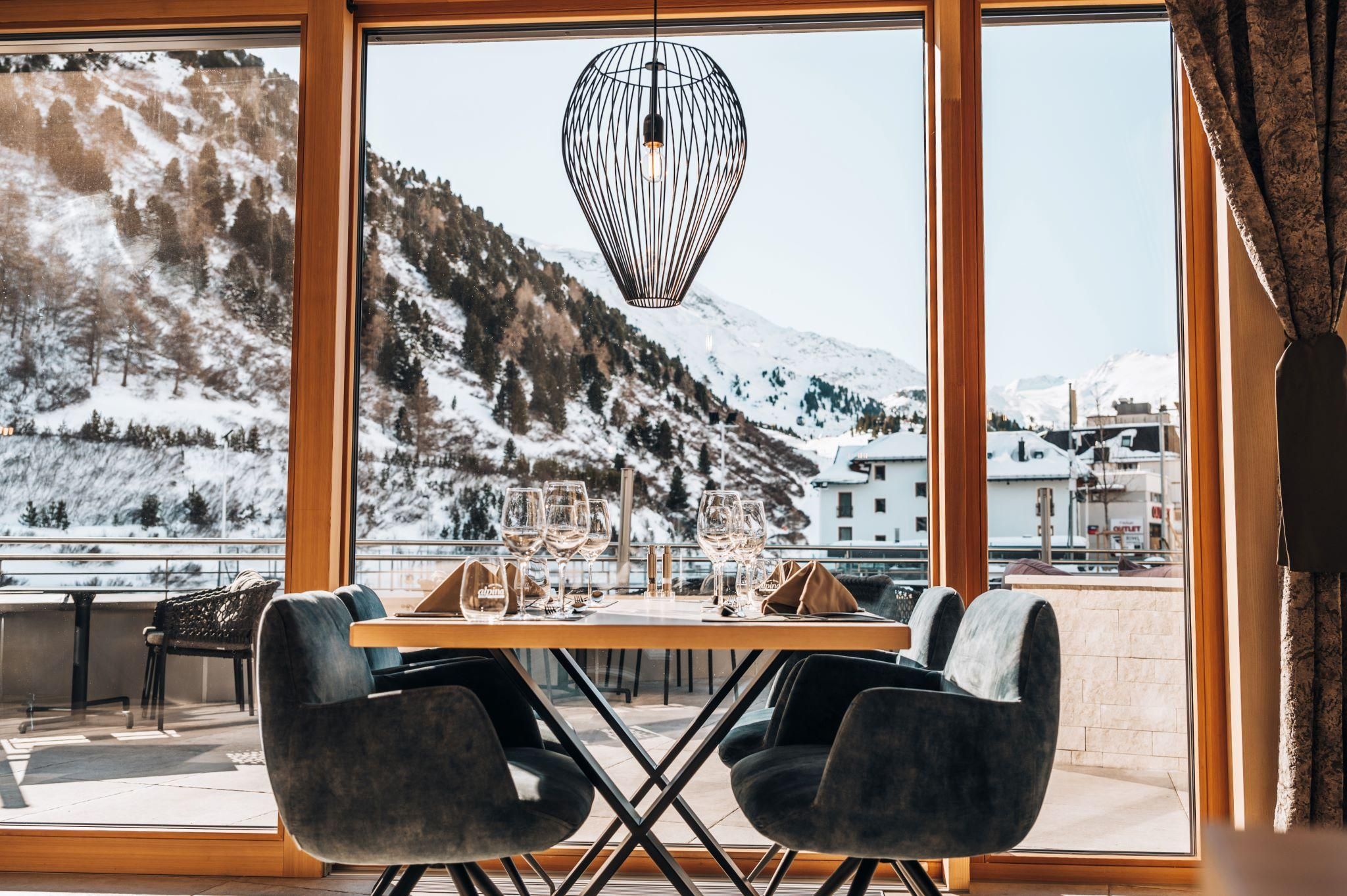 A modern restaurant with a table and chairs, overlooking snow-capped mountains. Large windows let in plenty of daylight.
