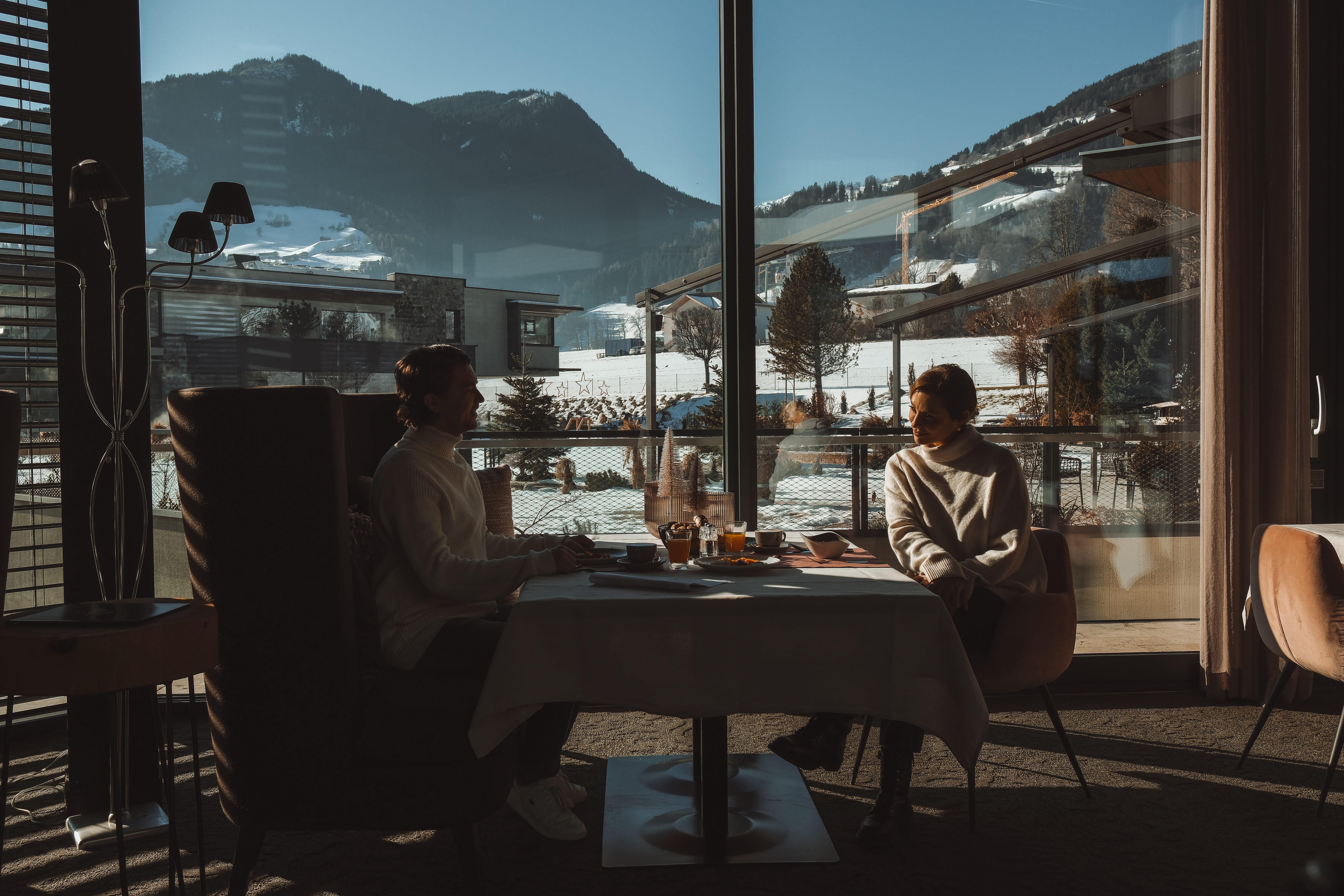 A couple is sitting at a table in a restaurant with a view of snow-covered mountains. The sun is shining, creating a cozy atmosphere.