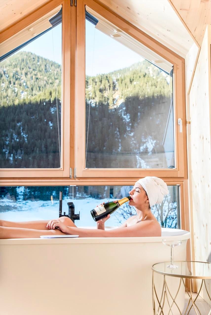 A relaxed person is sitting in a bathtub and enjoying champagne. In the background, there are mountains and a window with a view of nature.
