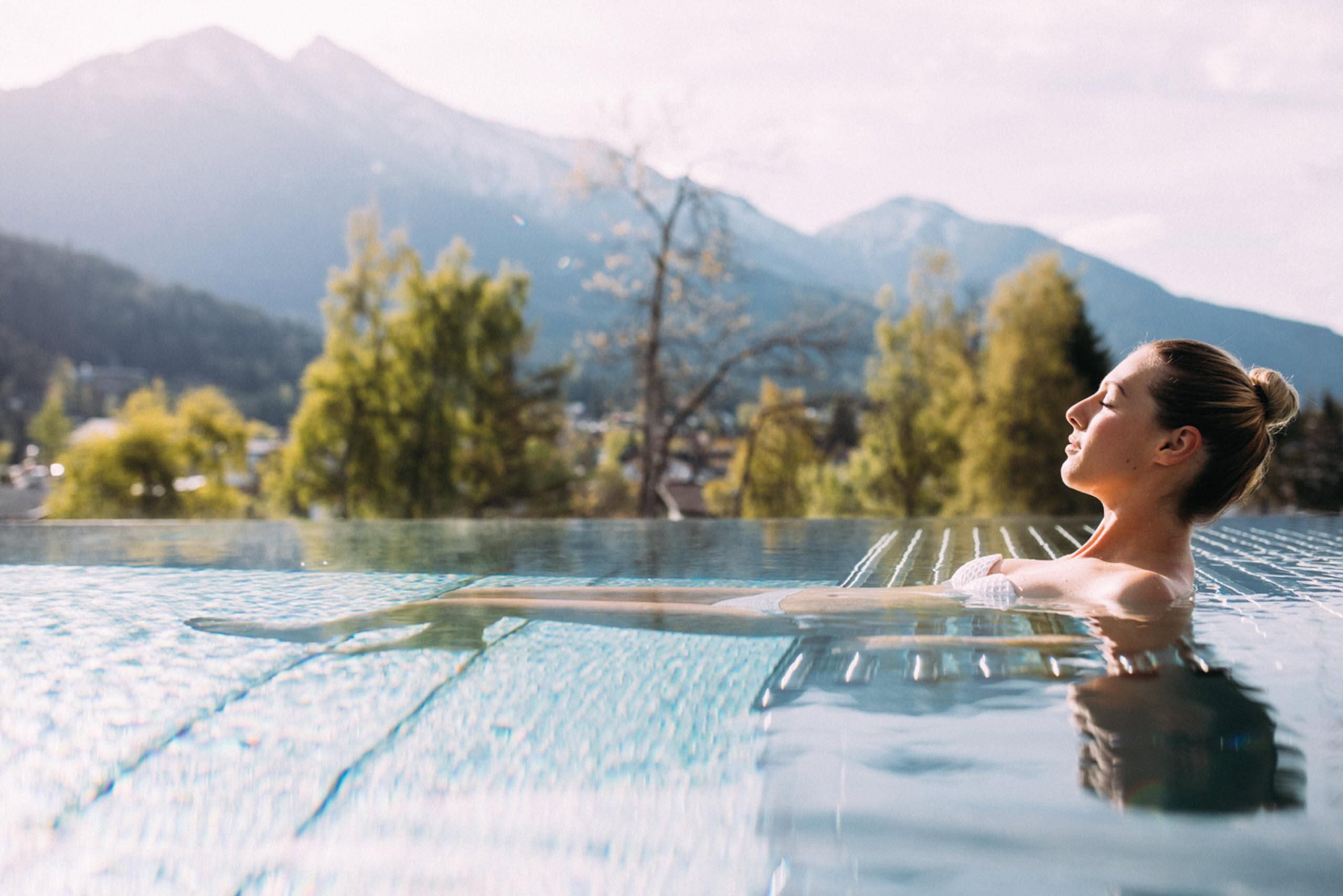 A woman relaxes in an infinity pool overlooking the mountains. The surroundings are green and sunny, creating a peaceful atmosphere.