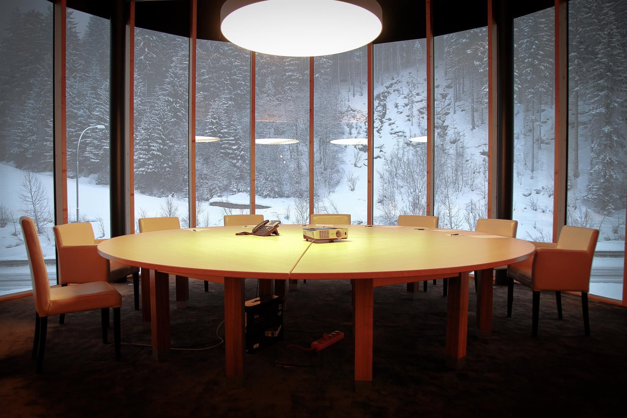 A modern conference room with a large, round table and chairs. Through the windows, a snow-covered landscape is visible.