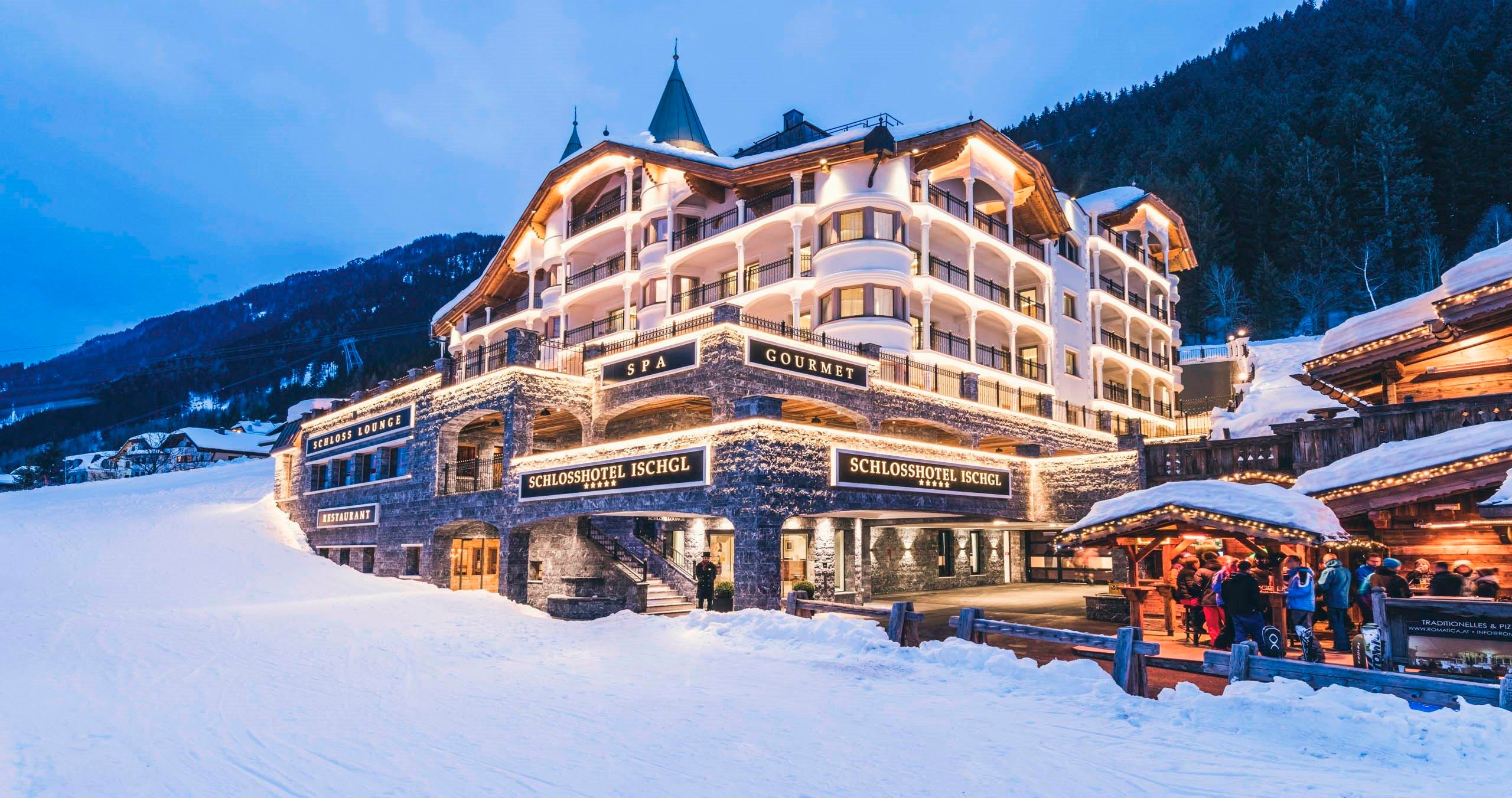 An elegant hotel in a snow-covered landscape, surrounded by mountains. The facade is illuminated and there is an inviting atmosphere.