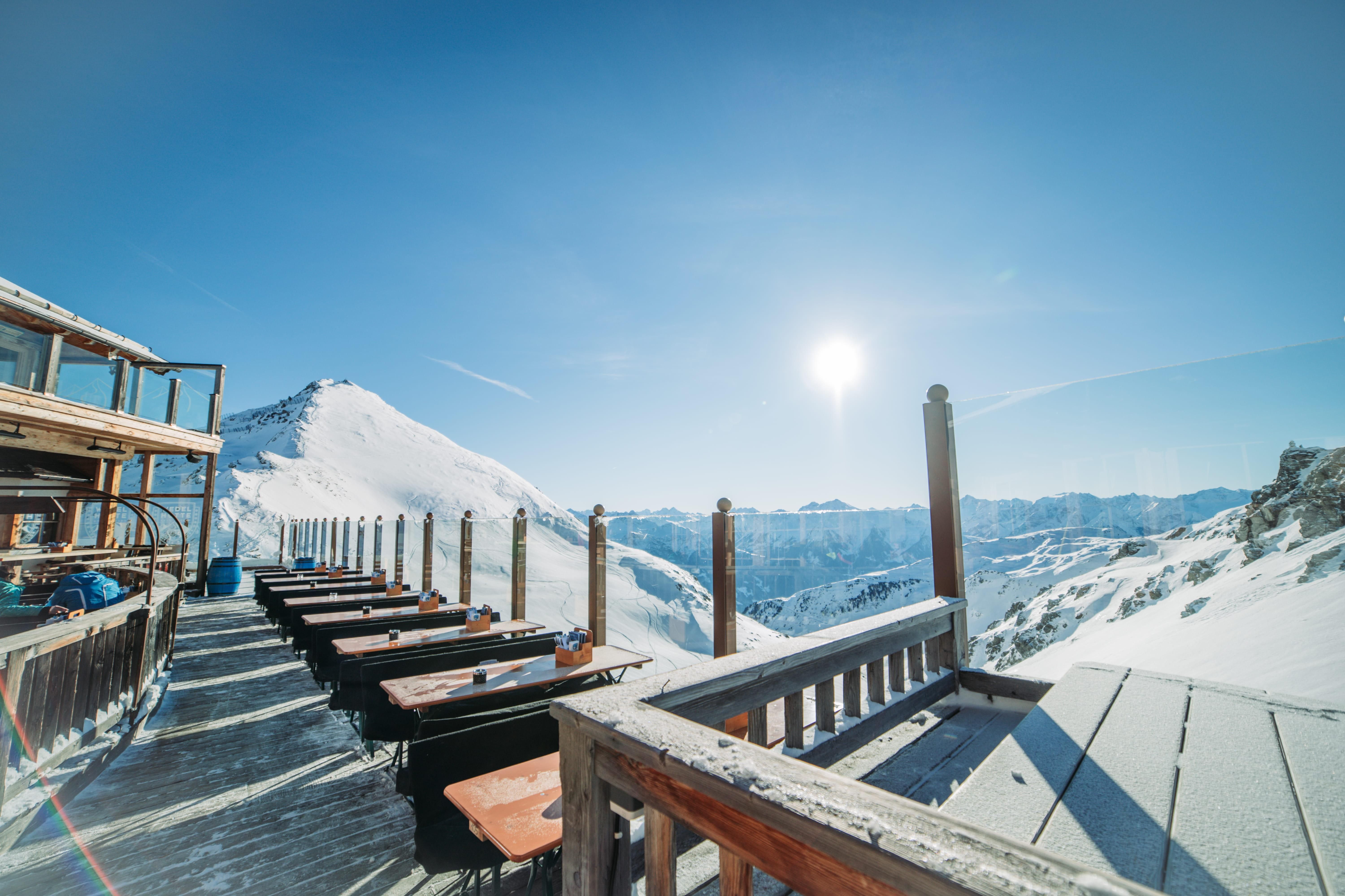 A sunny mountain landscape with snow-covered peaks and a terrace with tables. The clear sky shines over the alpine surroundings.