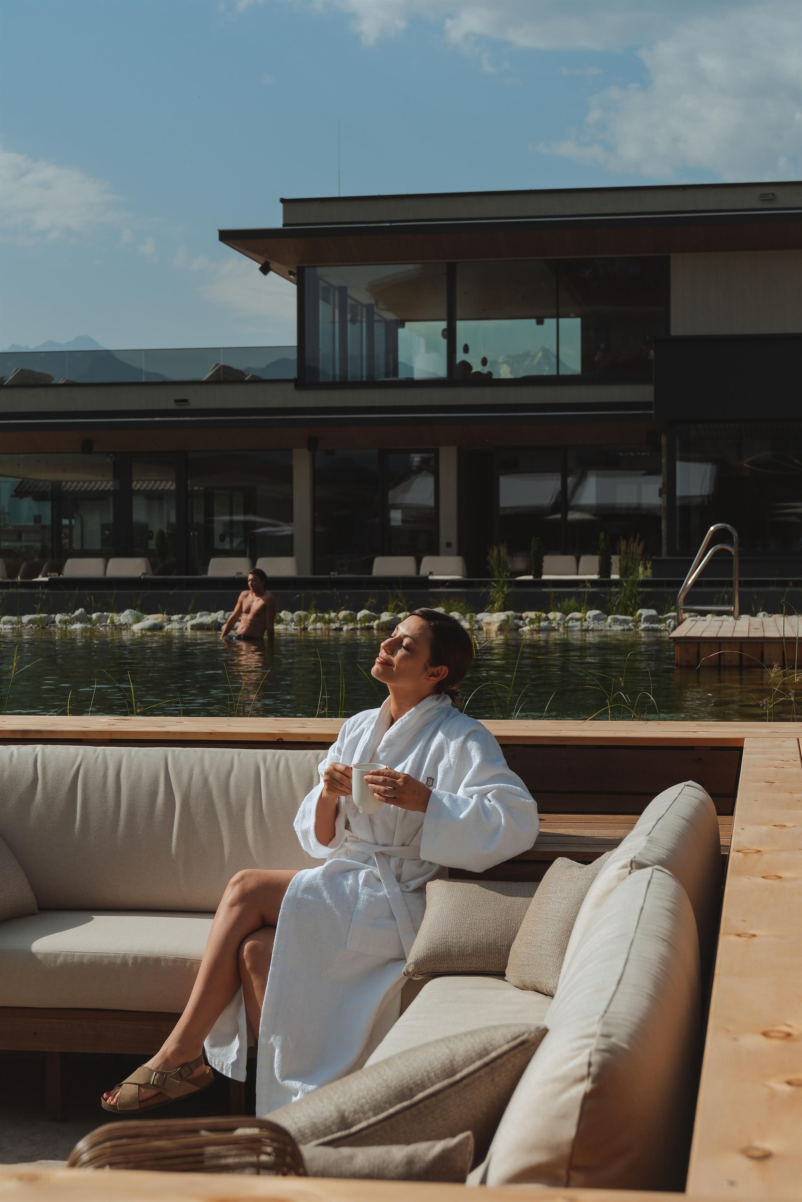 A woman in a white bathrobe is sitting relaxed on a couch next to a swimming pool. In the background, a modern building can be seen.