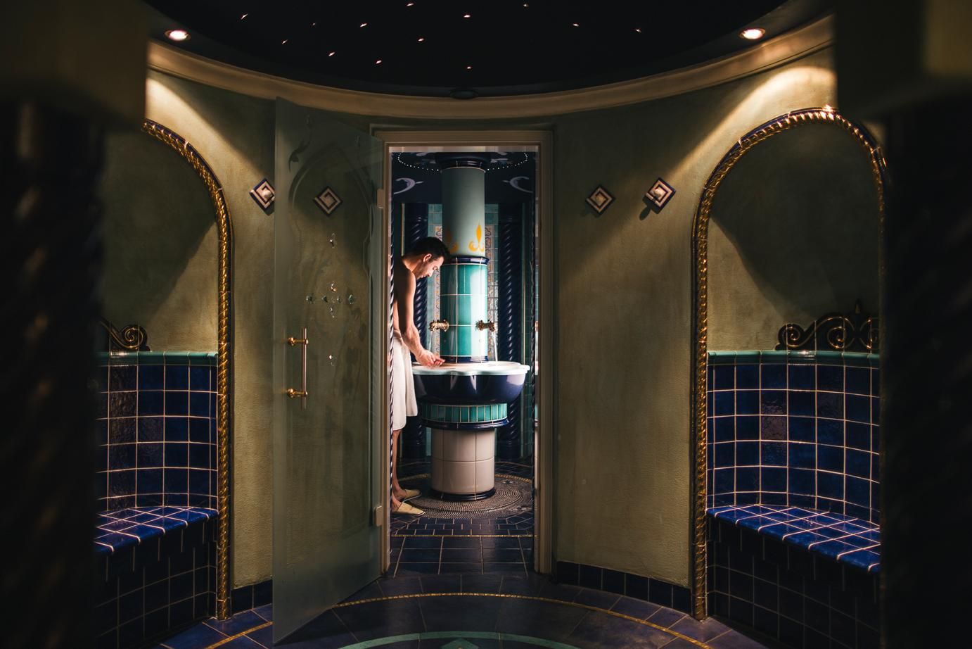An elegant bathroom with blue tiles and a central sink. A person stands at the sink and is experiencing a relaxing moment.