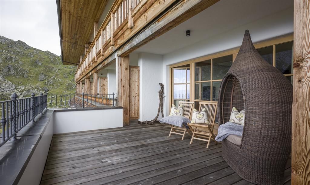 A cozy porch with a woven seat and two rocking chairs. In the background, green hills and a wooden building can be seen.