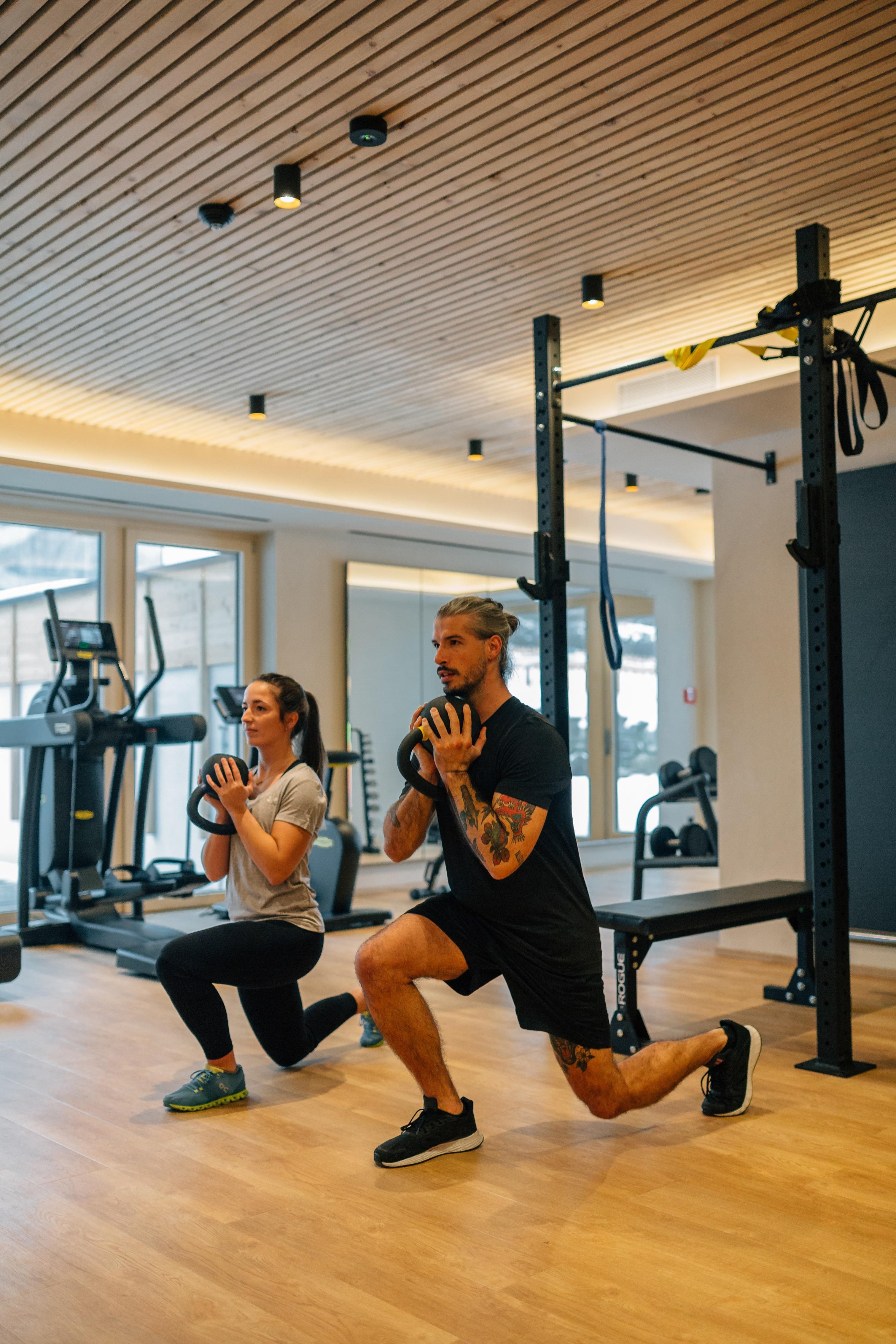 Two people are training in a modern gym. They are doing lunges with dumbbells.