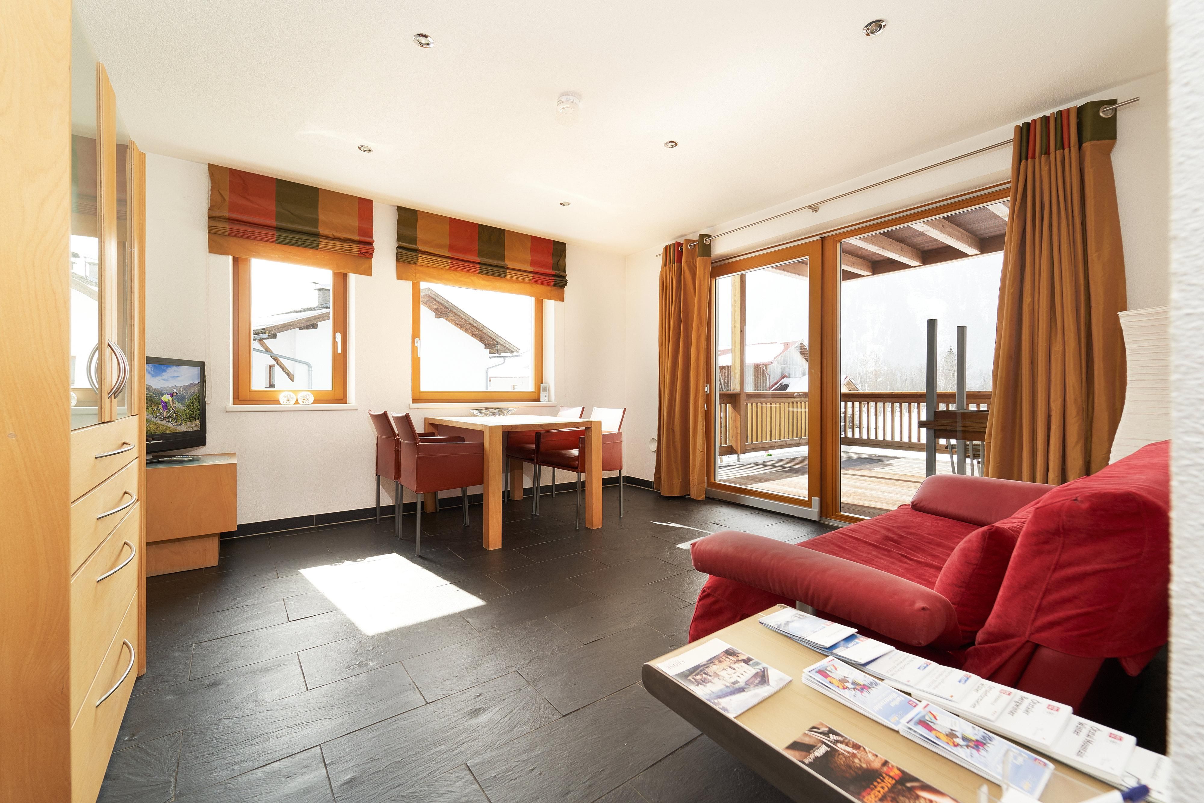 A bright living room with large windows and access to a balcony. The decor is modern and inviting with a red sofa and a dining area.